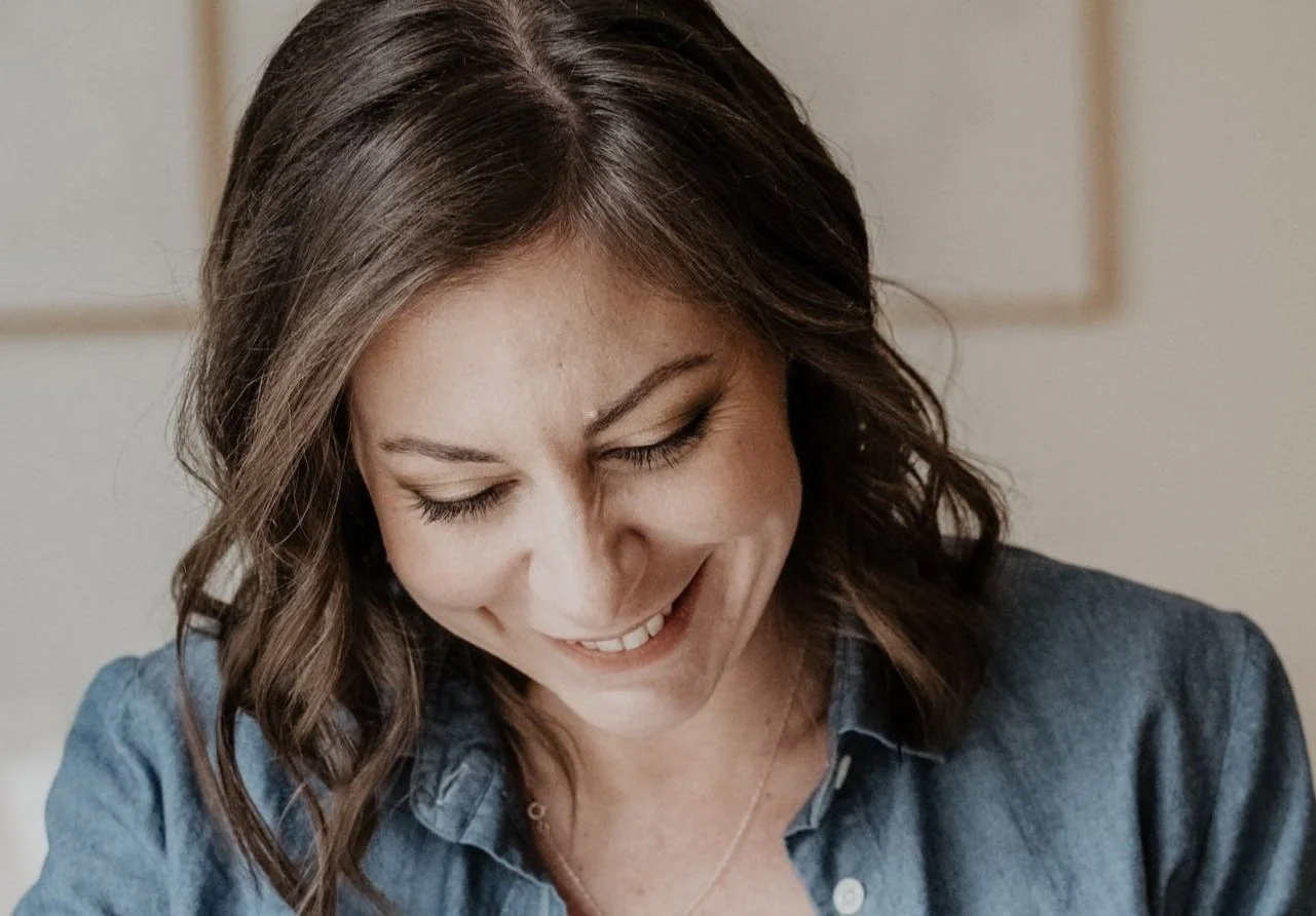 Eine lächelnde Frau mit braunen, gewellten Haaren trägt einen blauen Hemd und schaut nach unten.