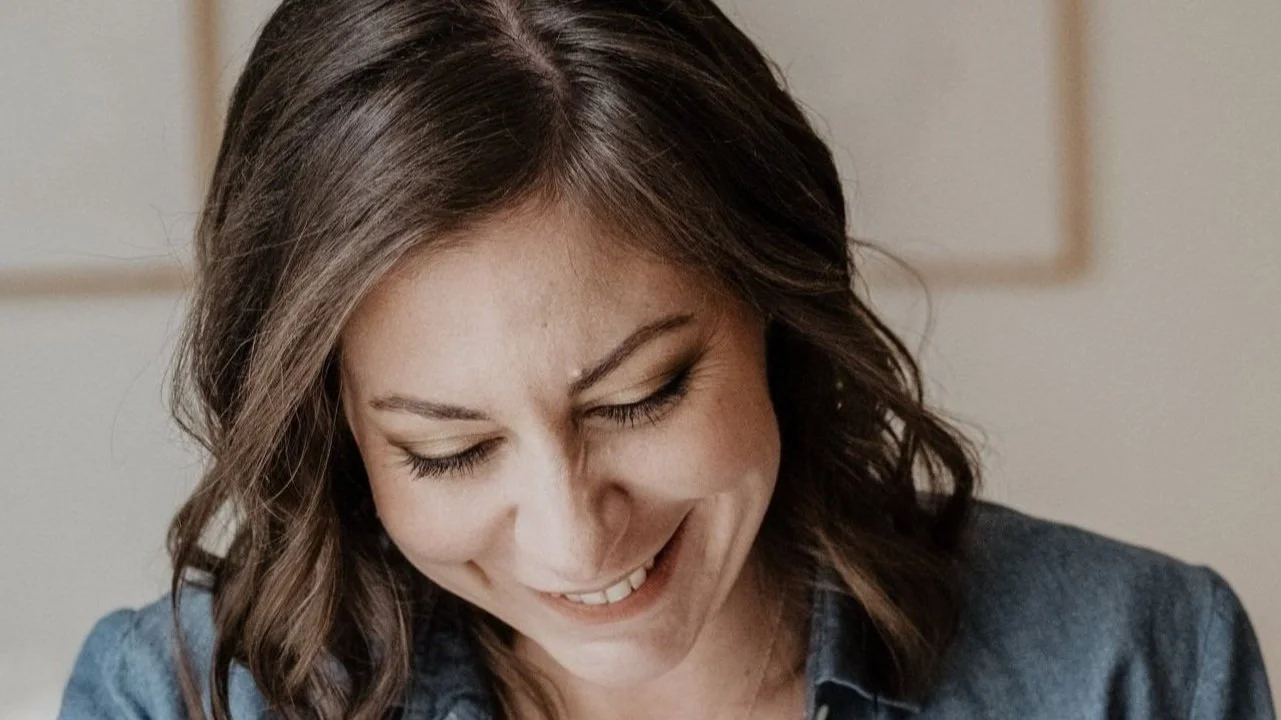 Lächelnde Frau mit braunen Haaren, die nach unten schaut, in einem blauen Hemd, im Innenraum.