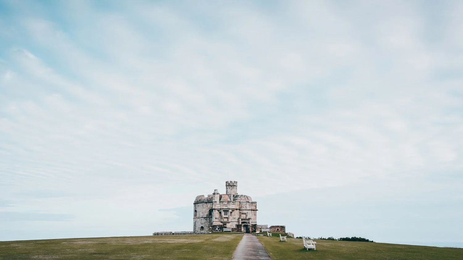 A historic stone castle on a grassy hill under a partly cloudy sky, with a pathway leading to the entrance and white benches along the side.