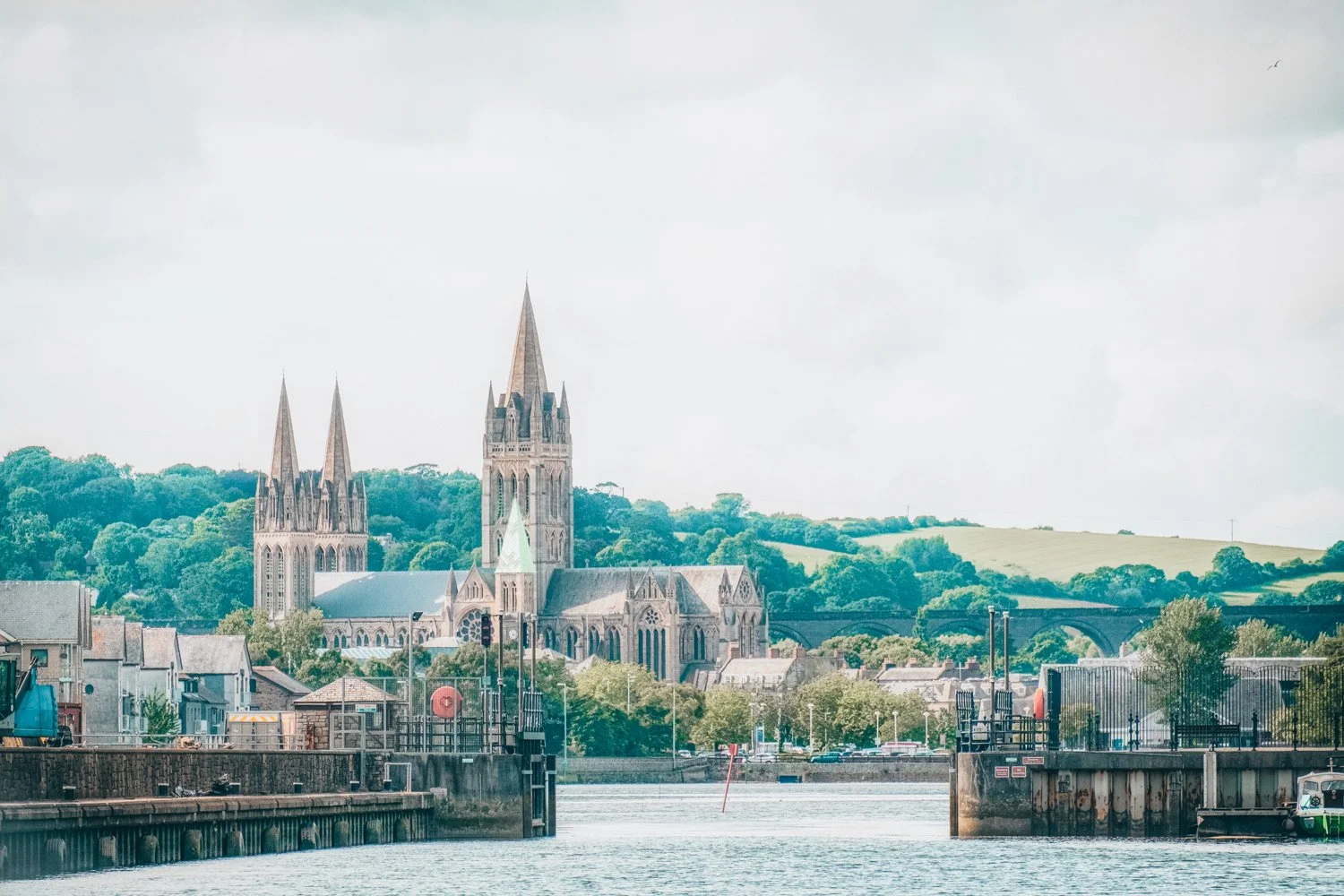 A scenic view of a historic church with tall spires across a body of water, with lush green hills in the background and cloudy sky overhead.
