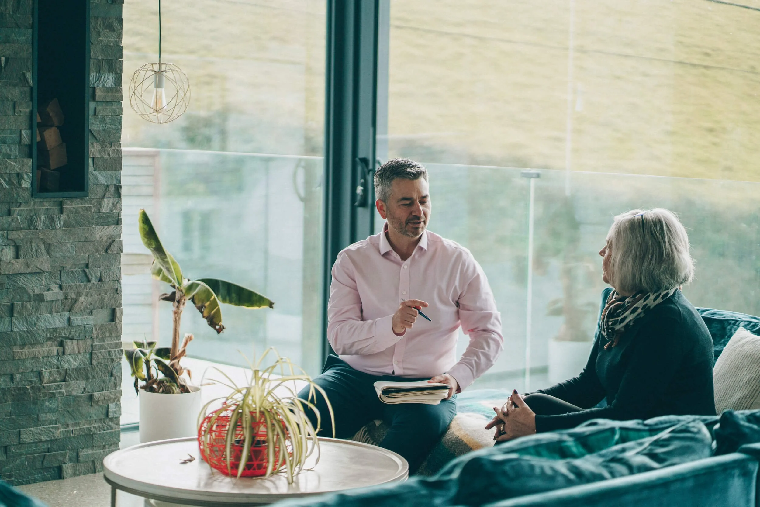 A man in a pink shirt and an older woman with white hair sitting and talking inside a modern living room with large glass windows, decorative plants, and a stone accent wall.
