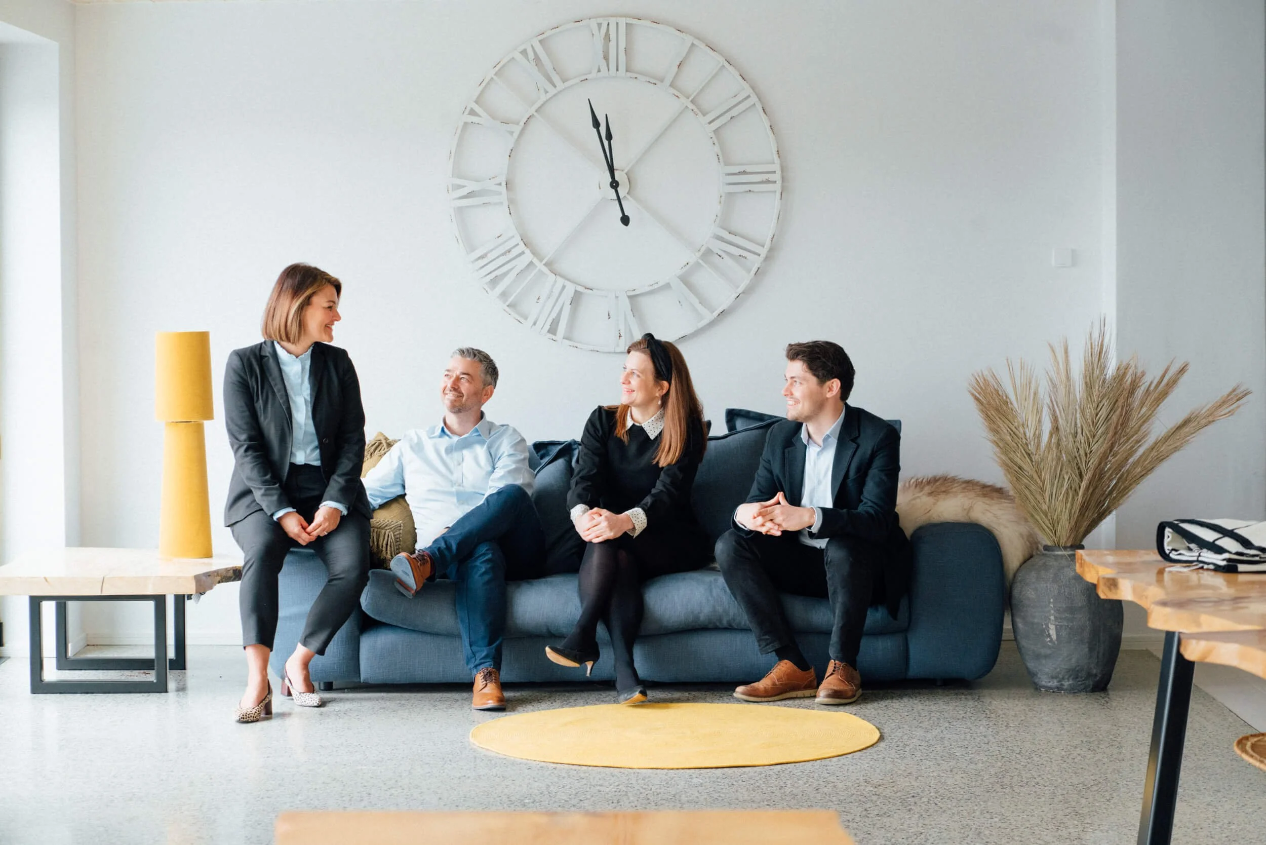 Four people in business attire sitting and standing in a modern living room, having a conversation. There is a large white wall clock behind them, a yellow lampshade on a side table, and a tall dried plant in a pot.