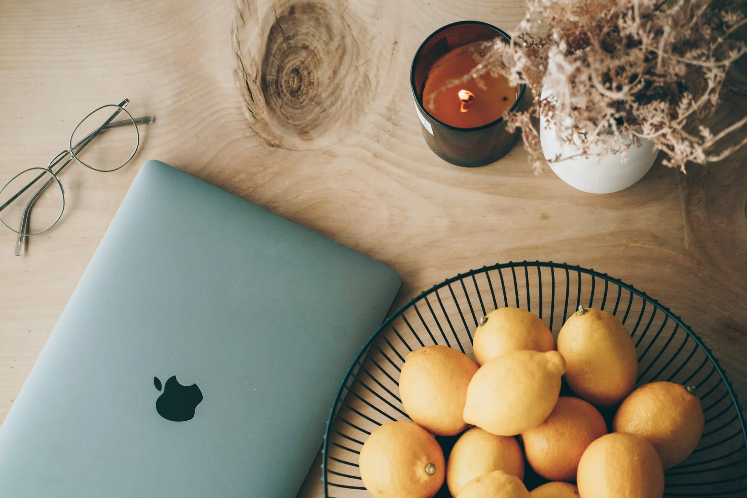 A laptop, sunglasses, a candle, a vase with dried flowers, and a basket of lemons on a wooden table.