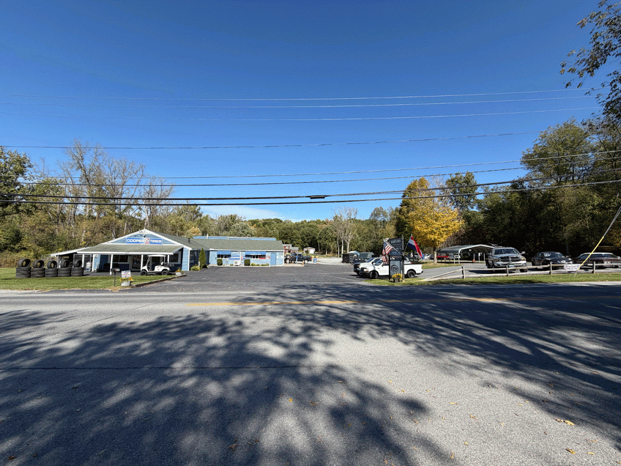 A parking lot with cars in front of a tire shop called Cooper Tires, surrounded by trees with some autumn foliage under a clear blue sky.