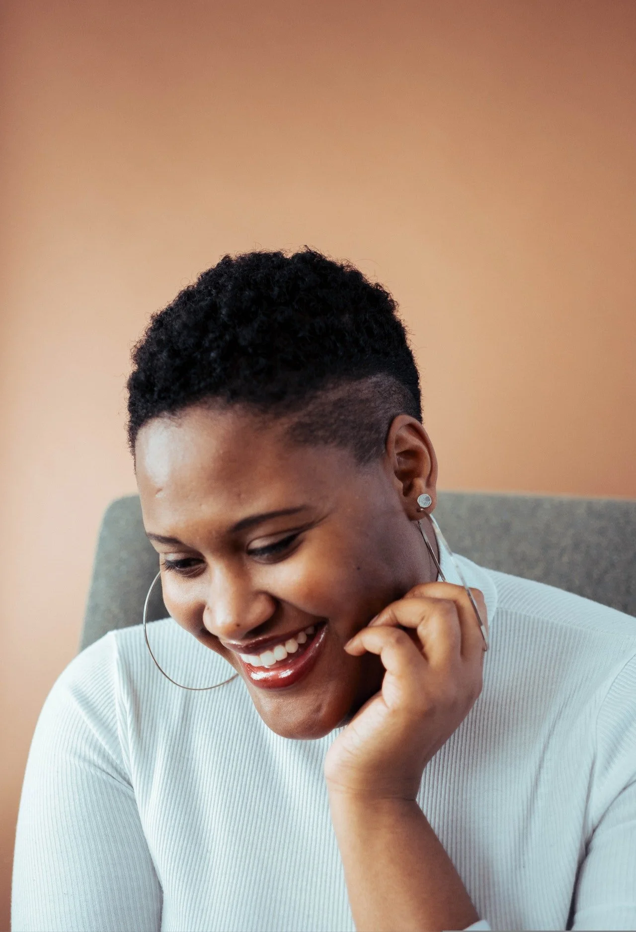 A black woman rests her hand on her chin, her large hoop earrings rest on top of her fingers, she smiles looking downward