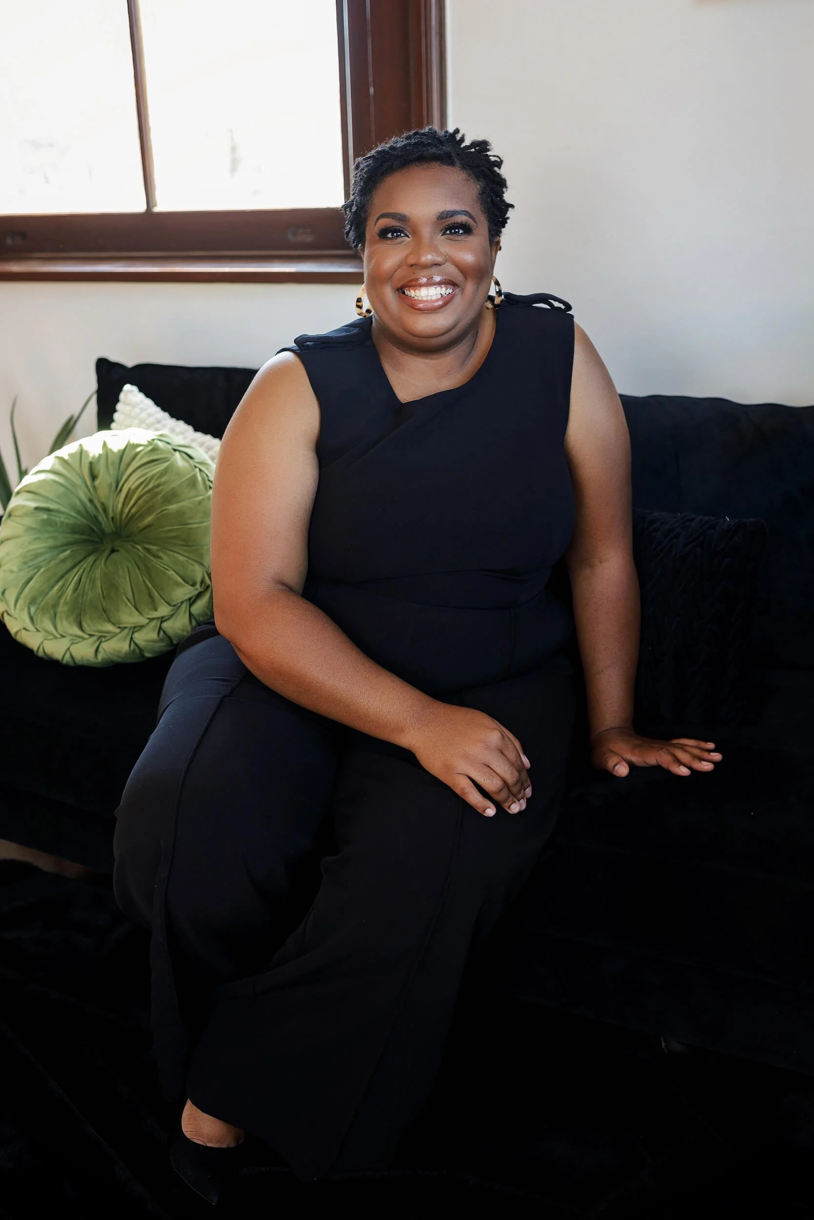 Dr Cecily Moore sits, smiling at the camera, as she leans one arm down on the couch below her, and one arm drapes across her lap