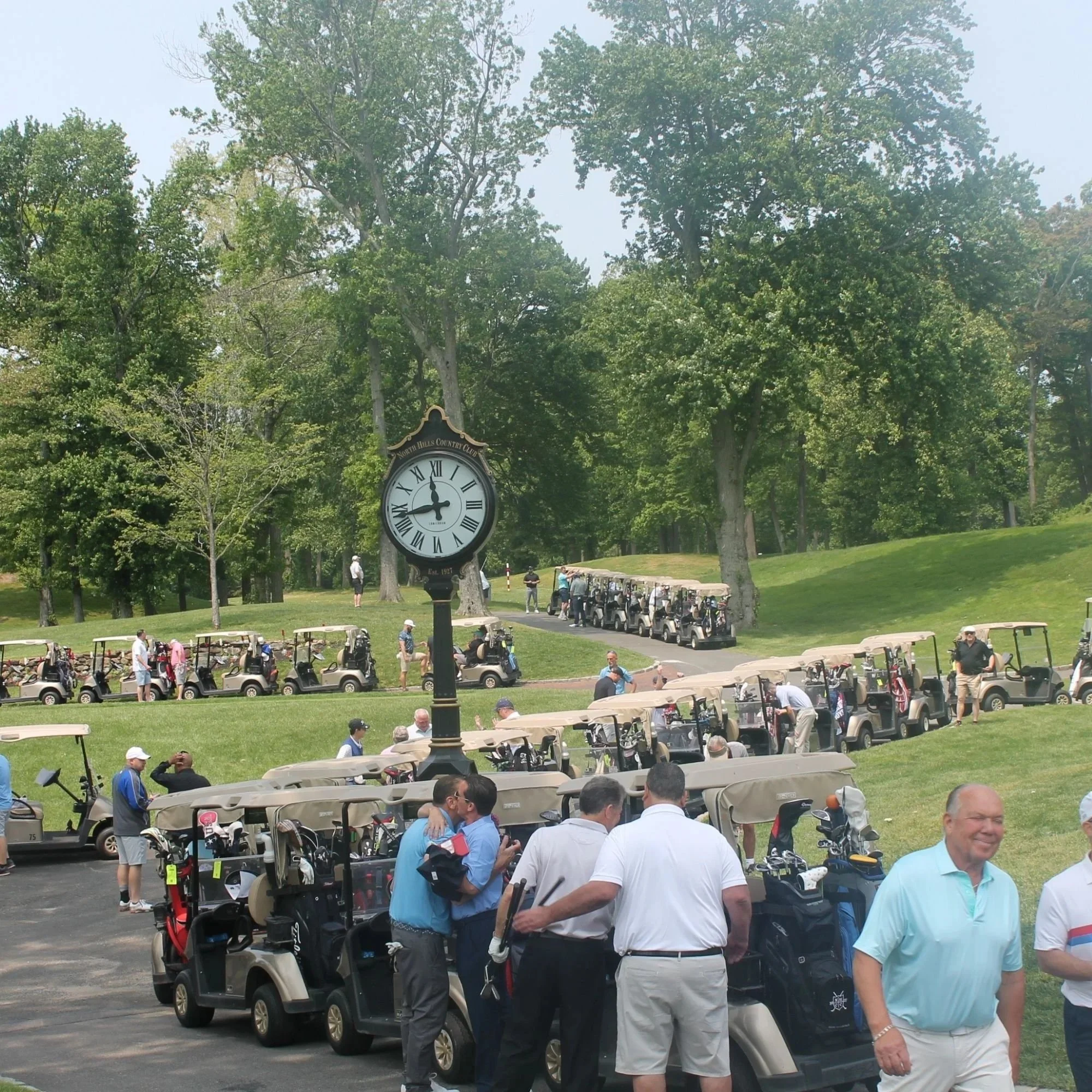 People preparing for a golf game at a golf course, with numerous golf carts lined up and a large clock showing approximately 12:03, surrounded by green trees.