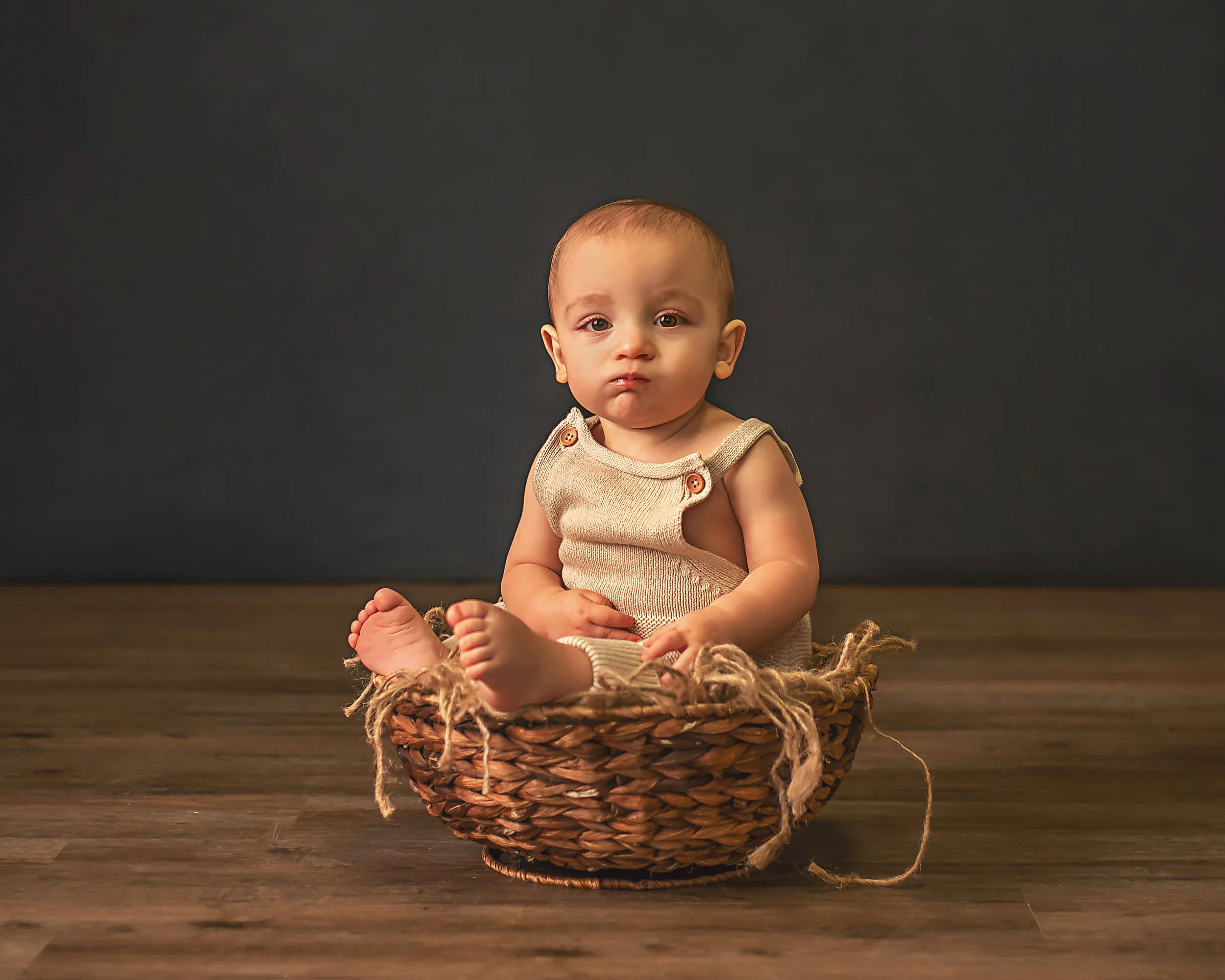 A baby sitting in a woven basket on a wooden floor against a dark background.