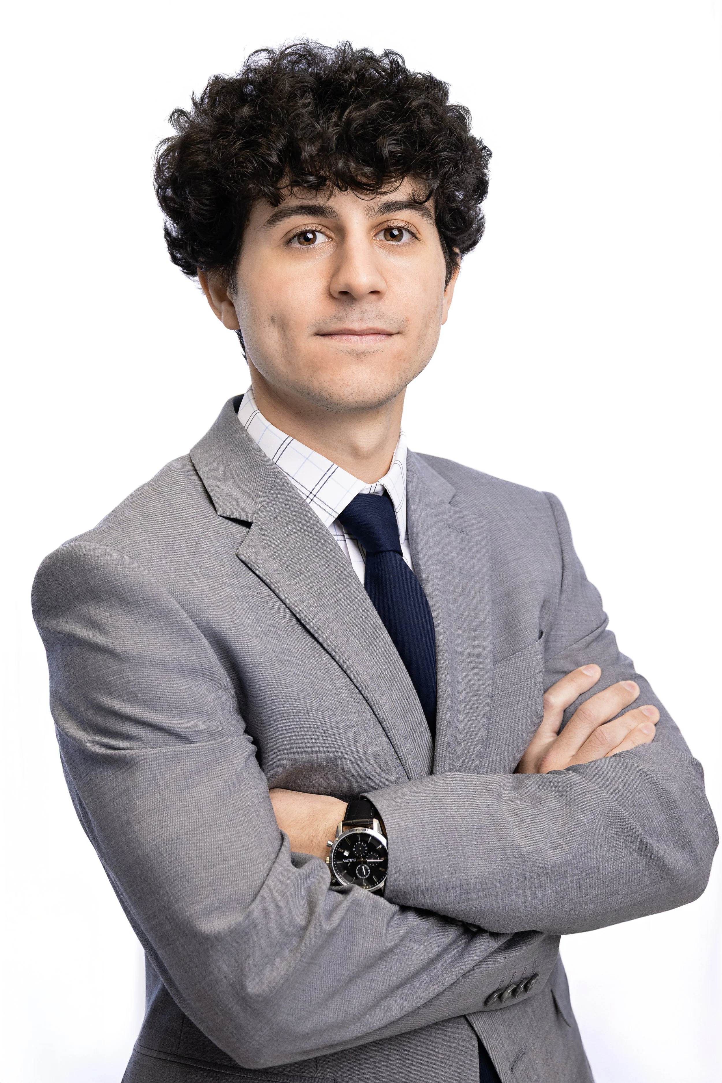 Professional man with curly dark hair wearing a gray suit, white shirt with plaid pattern, and a dark tie, standing with arms crossed against a white background.