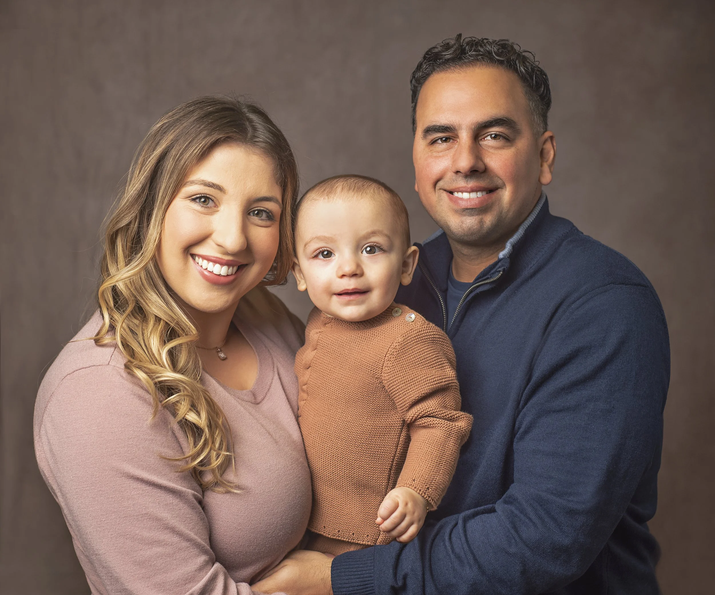 A happy family portrait featuring a woman with long blonde hair, a man with short dark hair, and a young child with short light hair, all smiling and looking at the camera, against a neutral background.