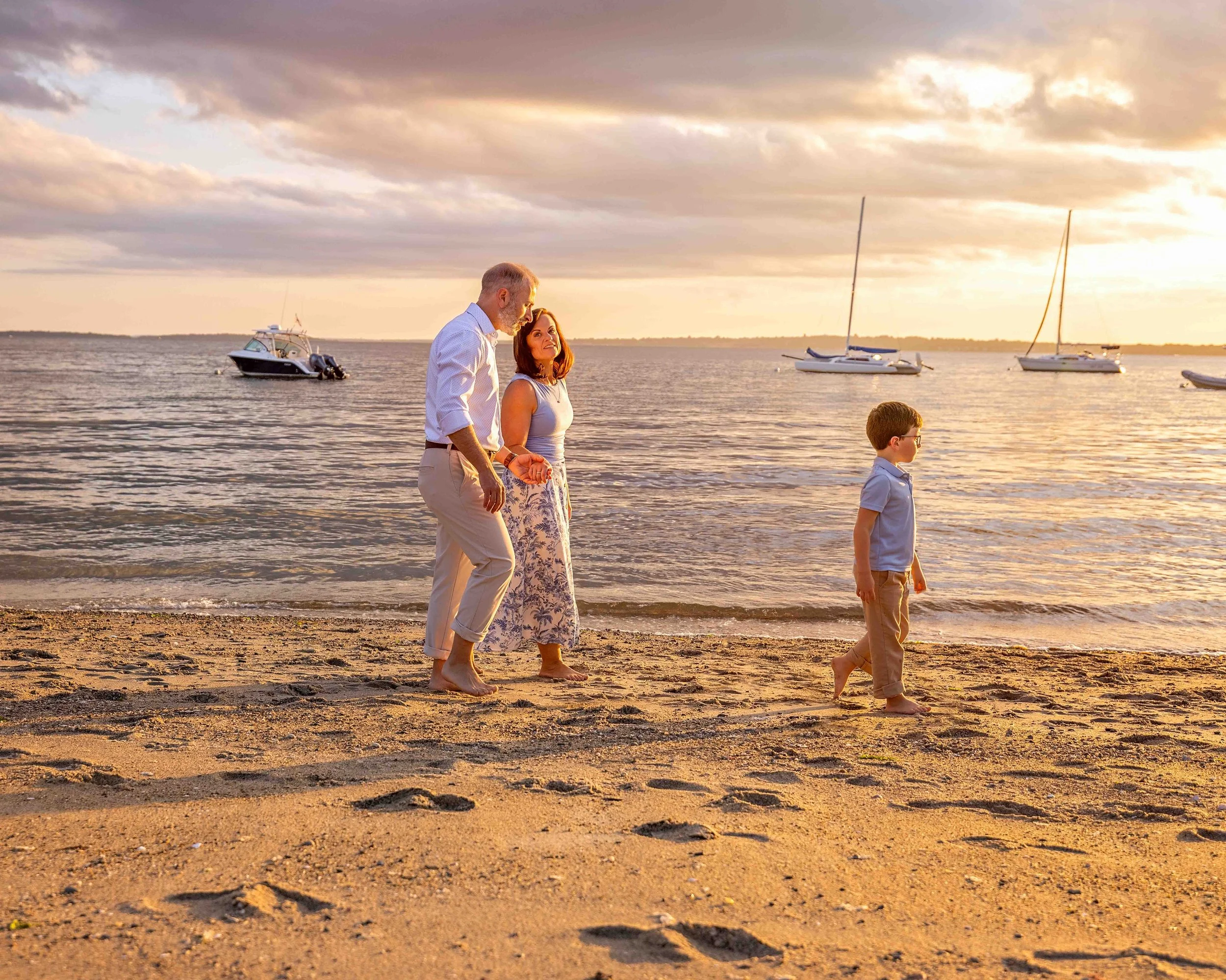 A family of three walking barefoot on a sandy beach at sunset, with sailboats anchored in the water in the background.