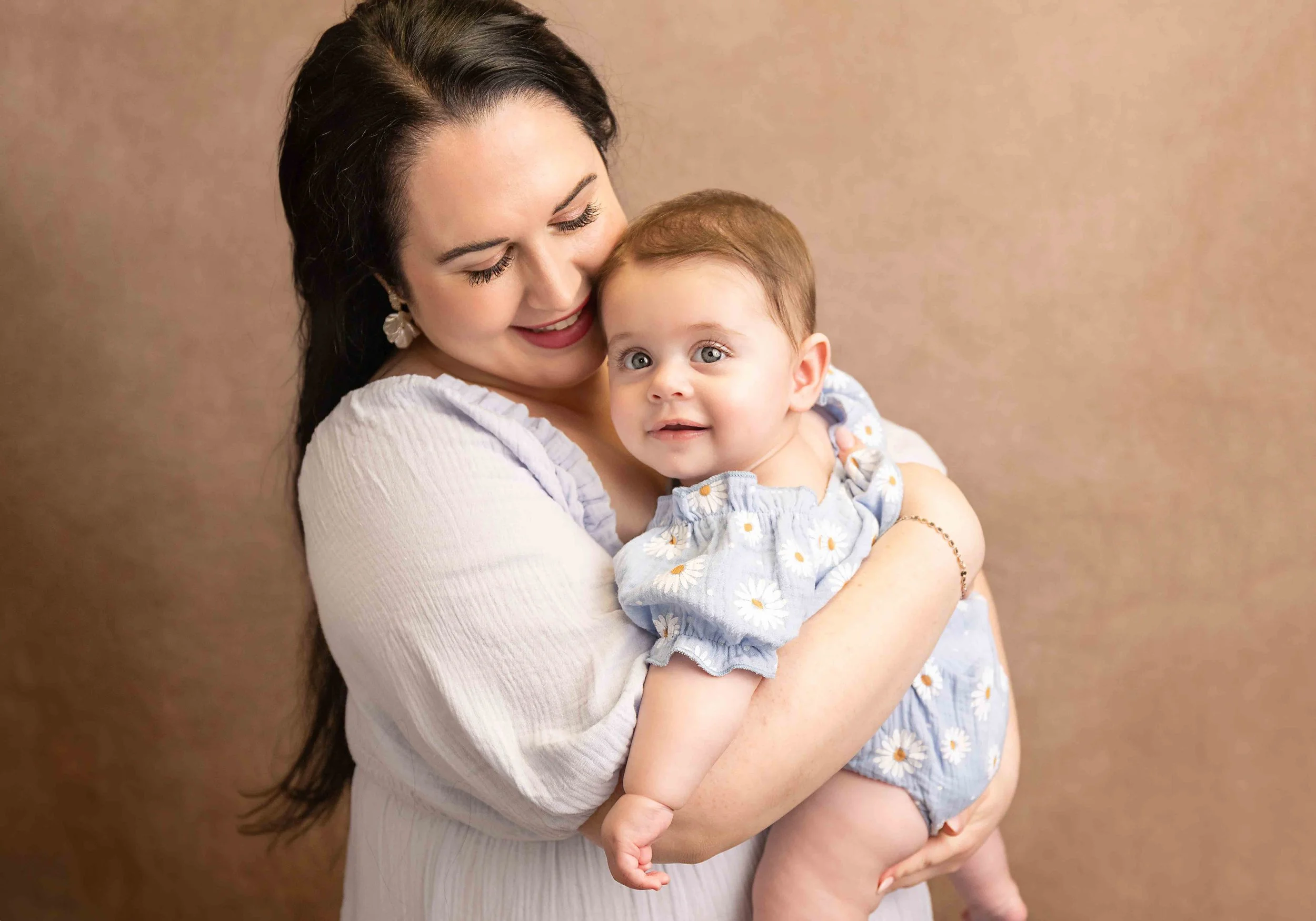 A woman with dark hair and earrings smiling and holding a baby girl with light hair and blue eyes, both facing the camera, against a plain background.