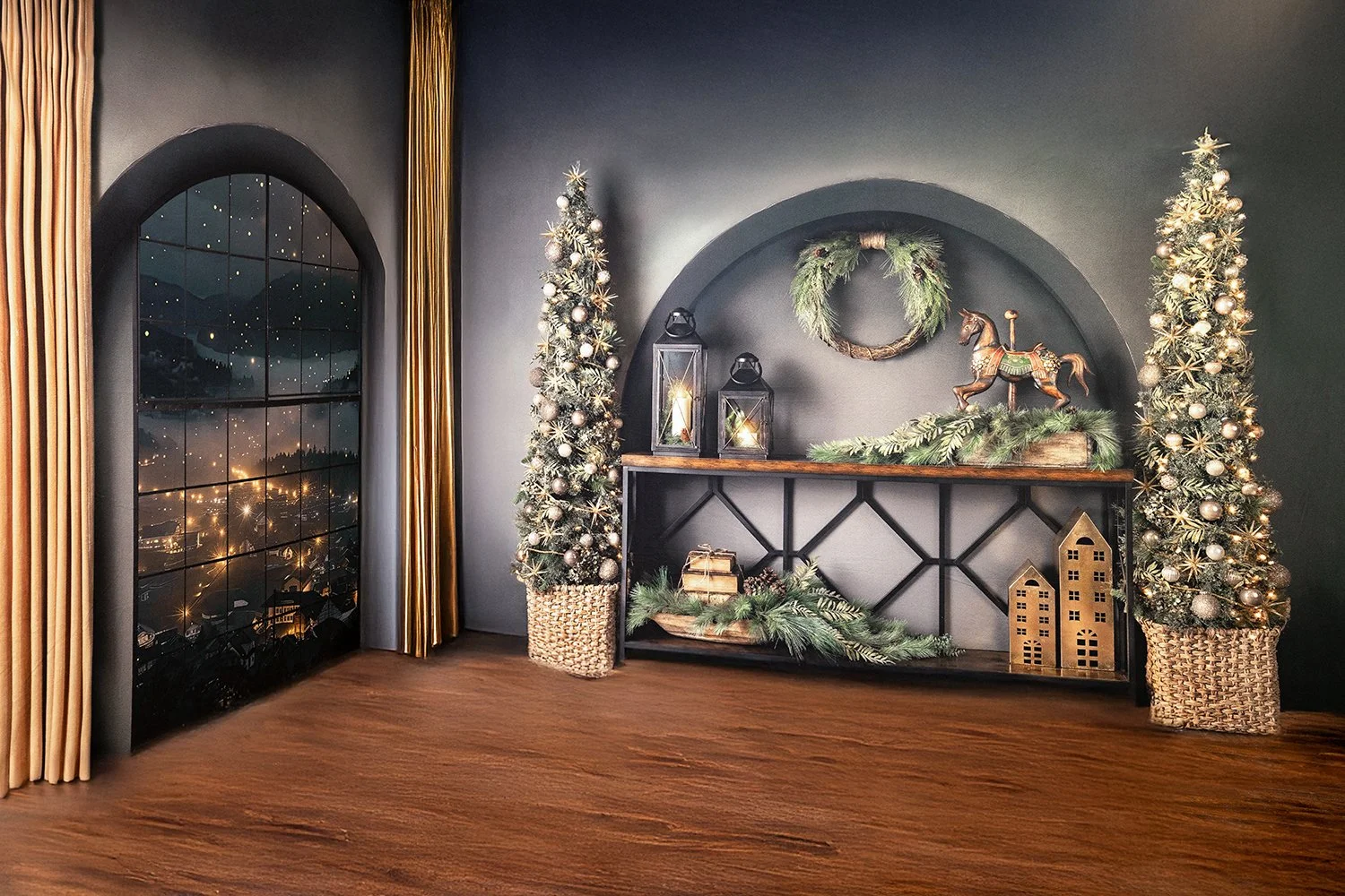Living room decorated for Christmas with two decorated Christmas trees, a wreath, lanterns with candles, a rocking horse, and golden houses on a console table, near a large window with golden curtains showing a night view of a town and mountains.