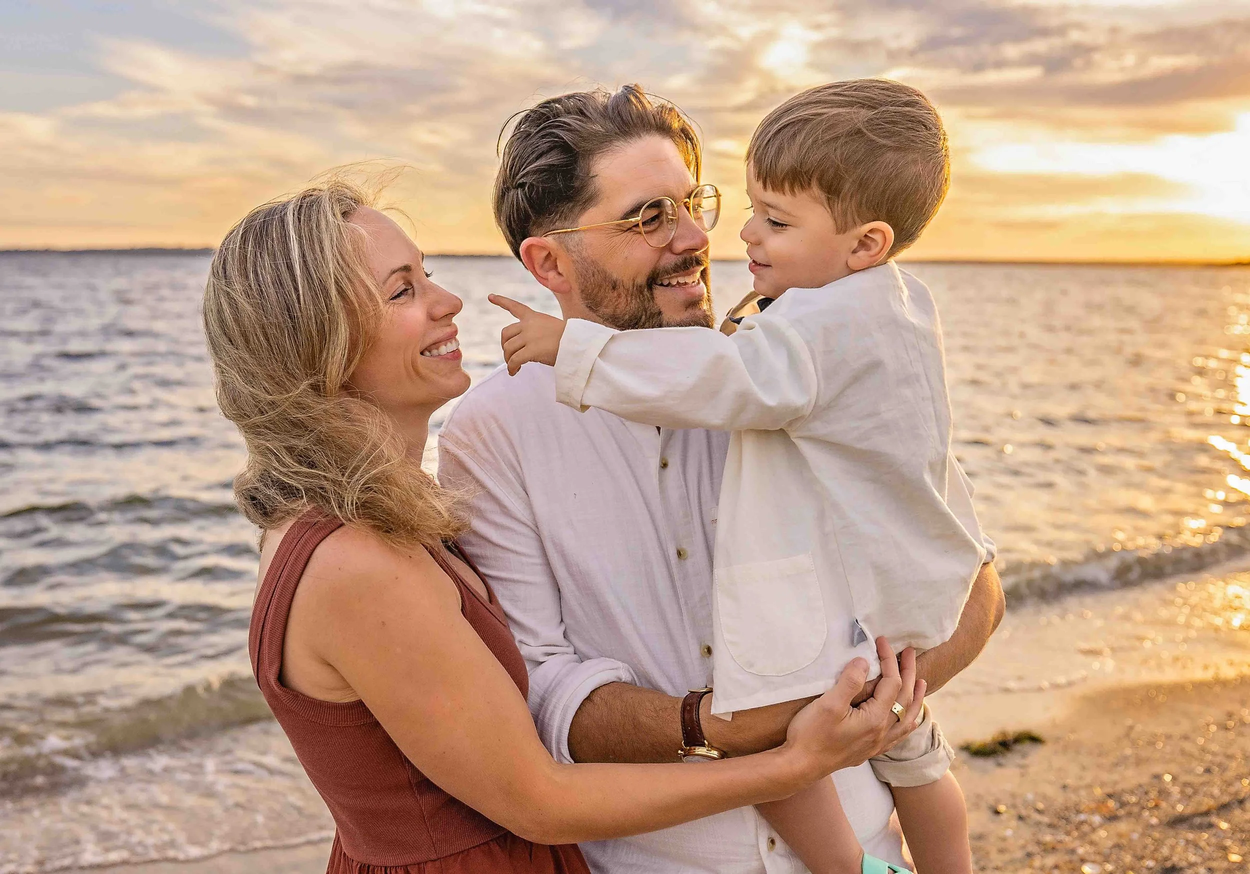 A family of three enjoying a sunset at the beach. The mother and father are holding their young son, who is pointing at them, with all three smiling.