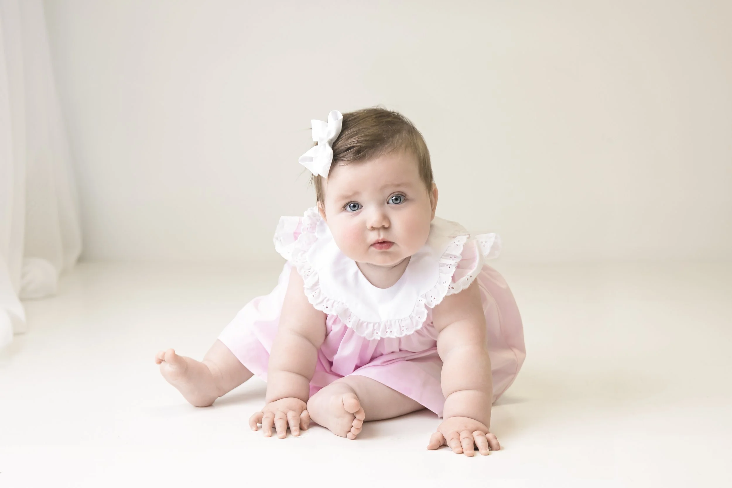 Adorable baby girl with blue eyes and brown hair, wearing a pink dress with white ruffled collar and bow, sitting on the floor against a plain light background.