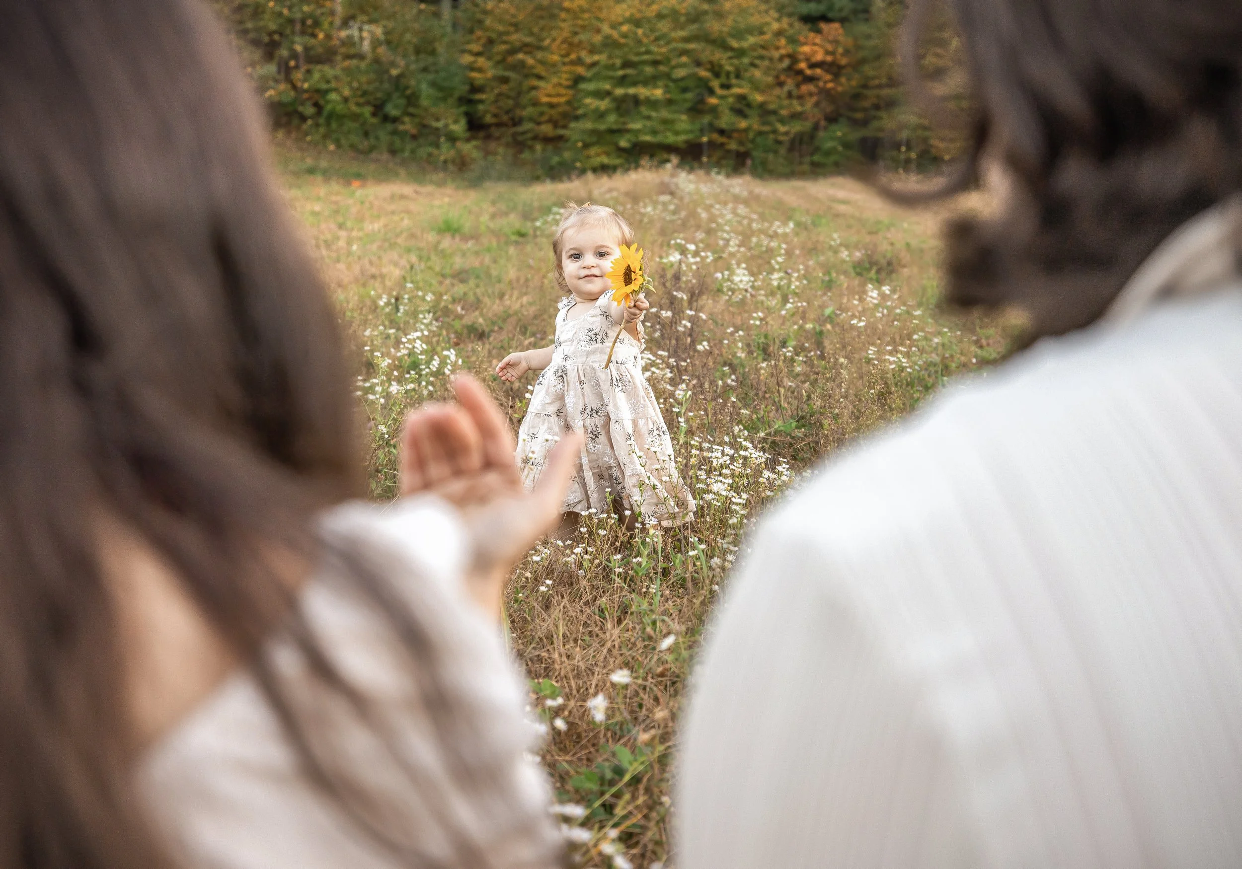 A young girl in a floral dress holding a sunflower, standing in a field of wildflowers with two women partially visible in the foreground, outdoors during fall.