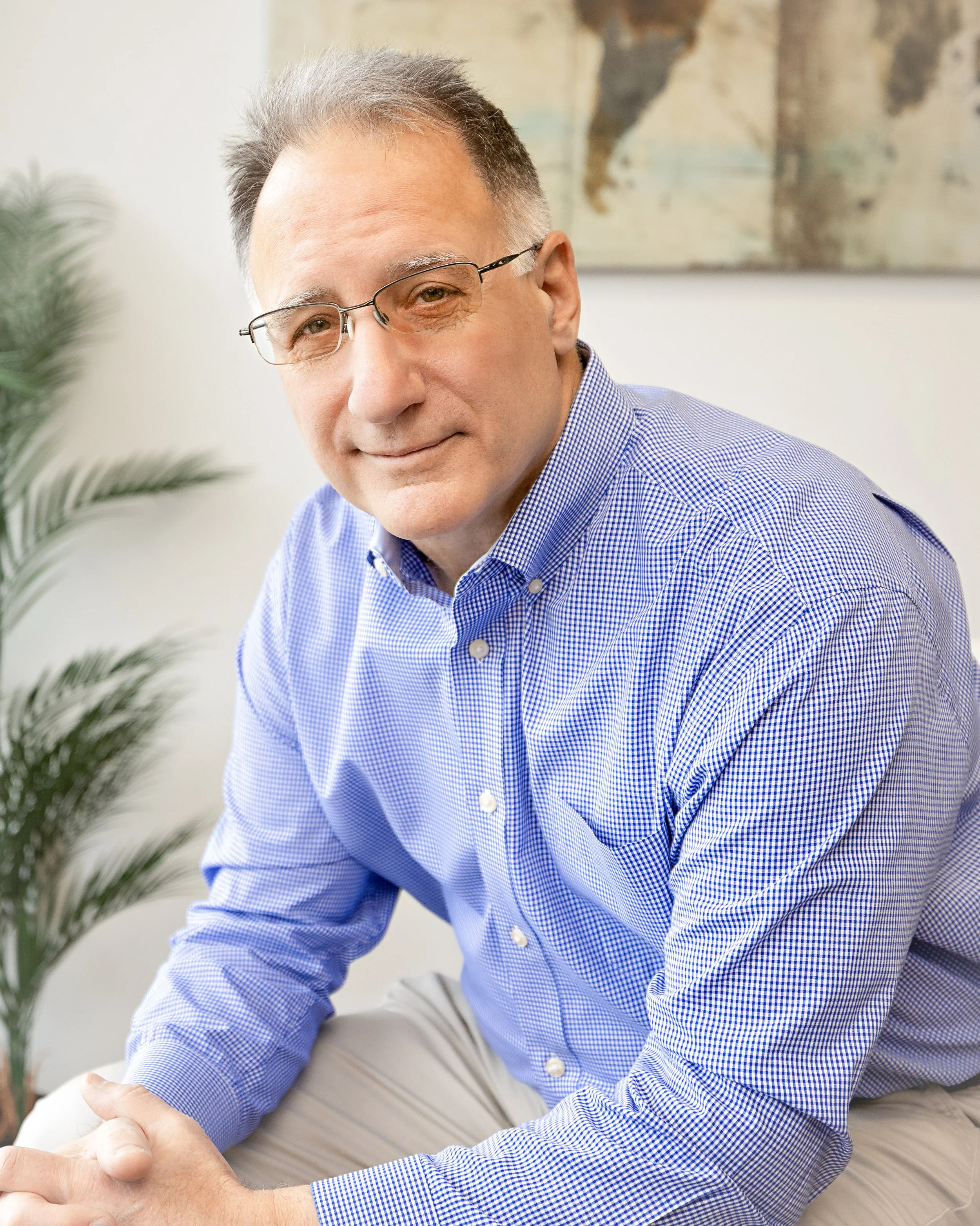 A middle-aged man with glasses and gray hair, wearing a blue checkered shirt, is sitting indoors near a plant, with a world map on the wall behind him.