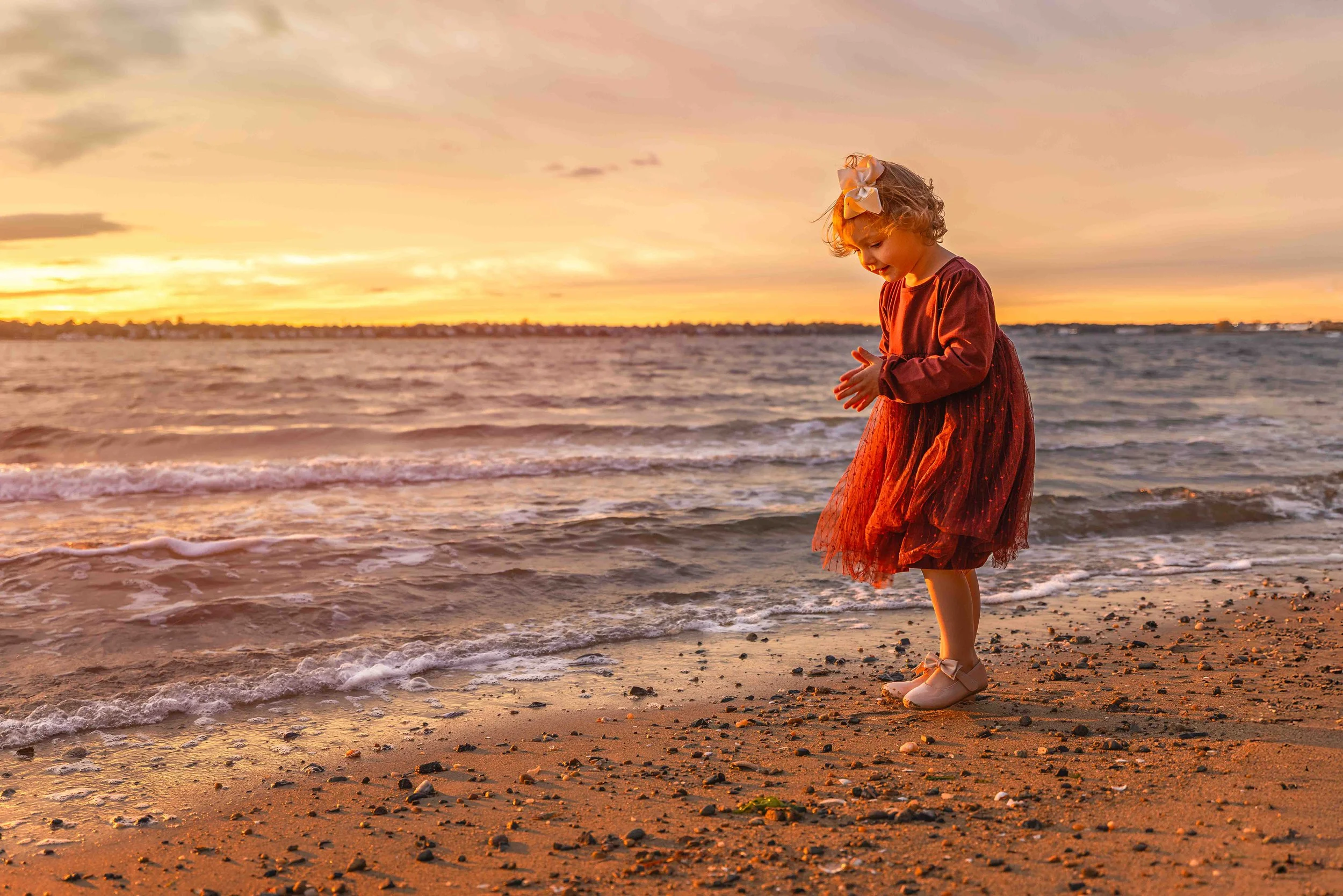 A young girl in a red dress with bows in her hair standing on a sandy beach at sunset, looking down at the water.