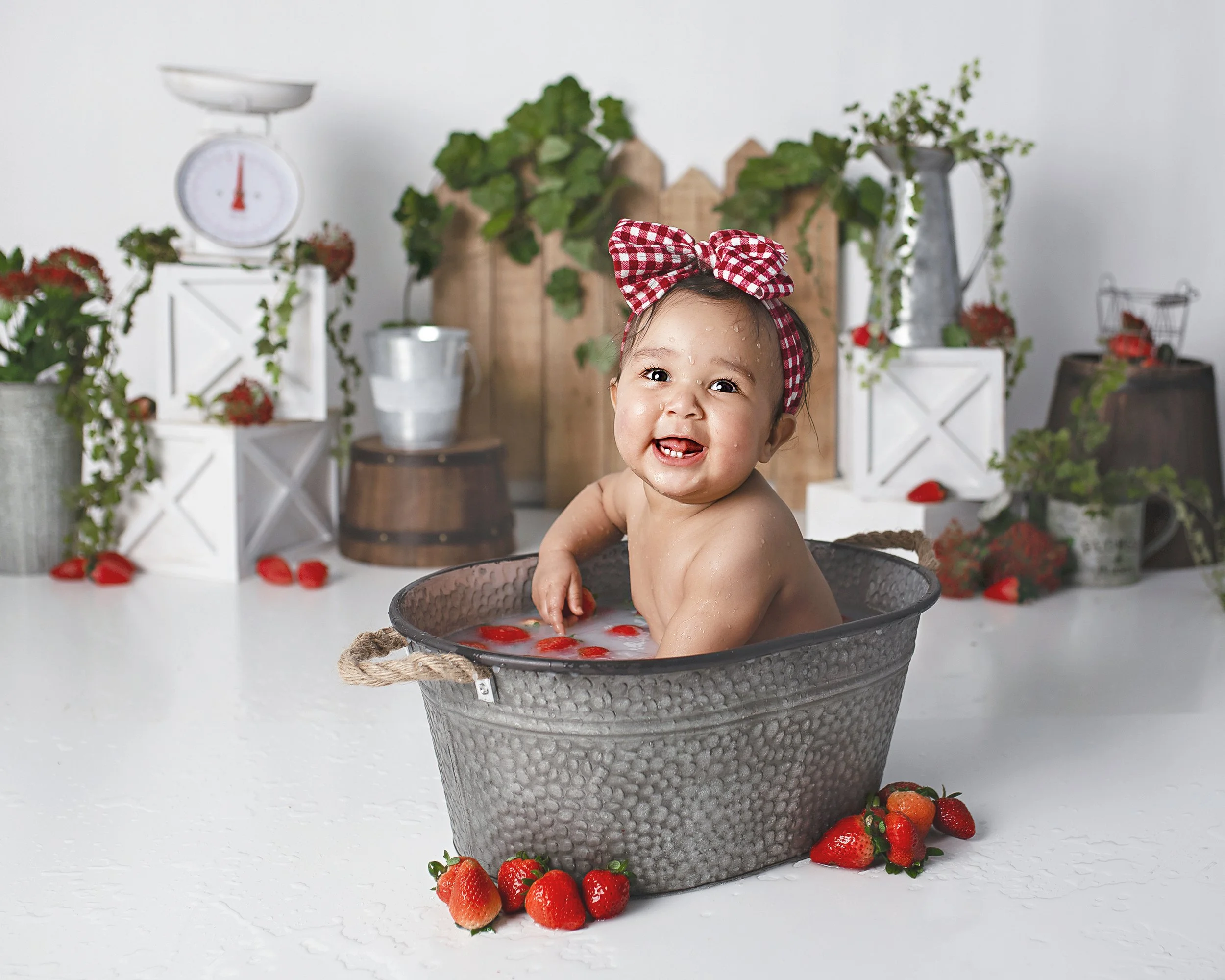A baby with a red and white checkered bow in hair sitting in a gray tub filled with water and strawberries, smiling at the camera in a bright room decorated with strawberries and farm-themed decor.
