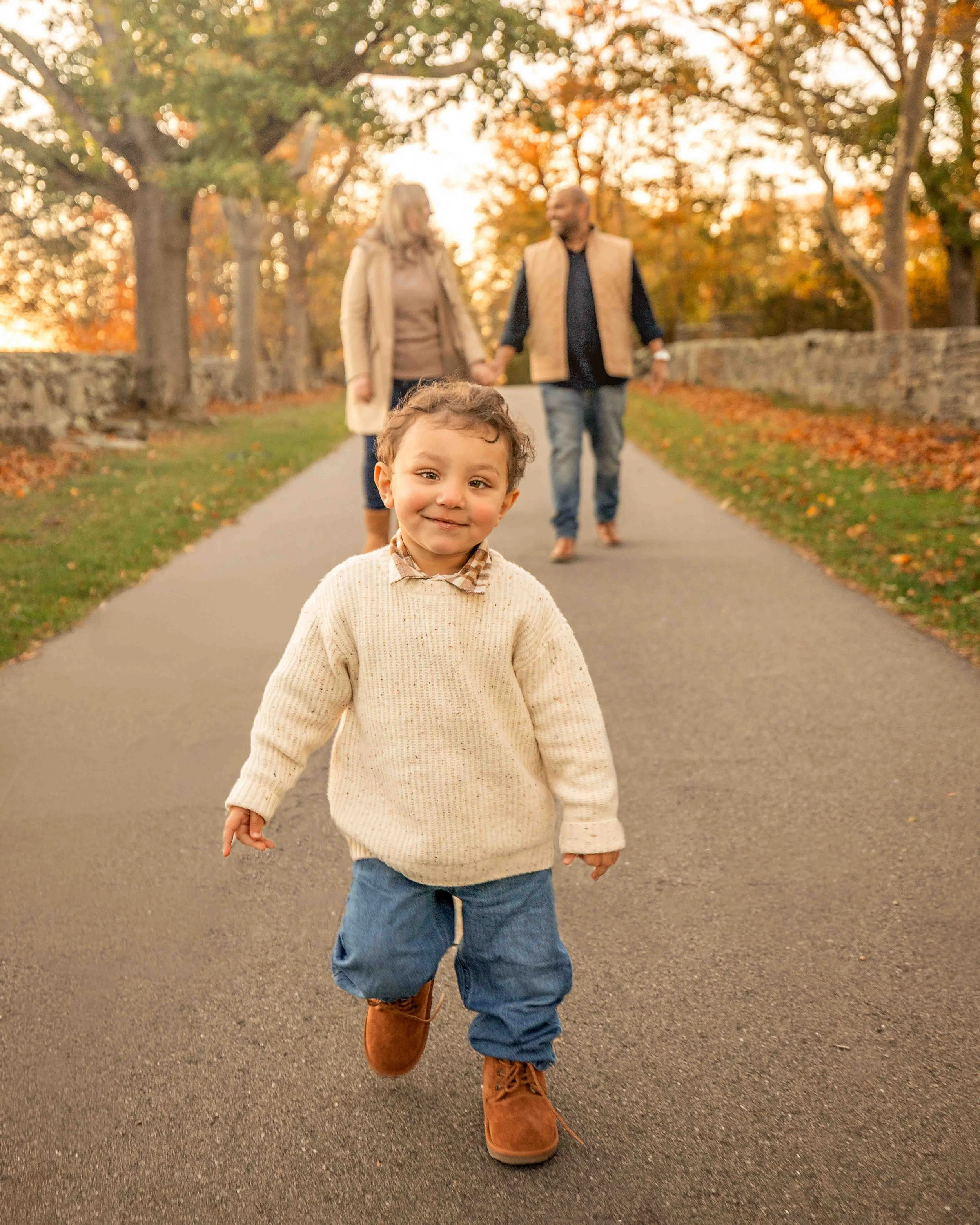 A young child with curly hair, wearing a cream sweater, jeans, and brown boots, runs on a paved path in an autumn park. In the background, a couple holding hands walks behind the child, surrounded by trees with fall foliage.