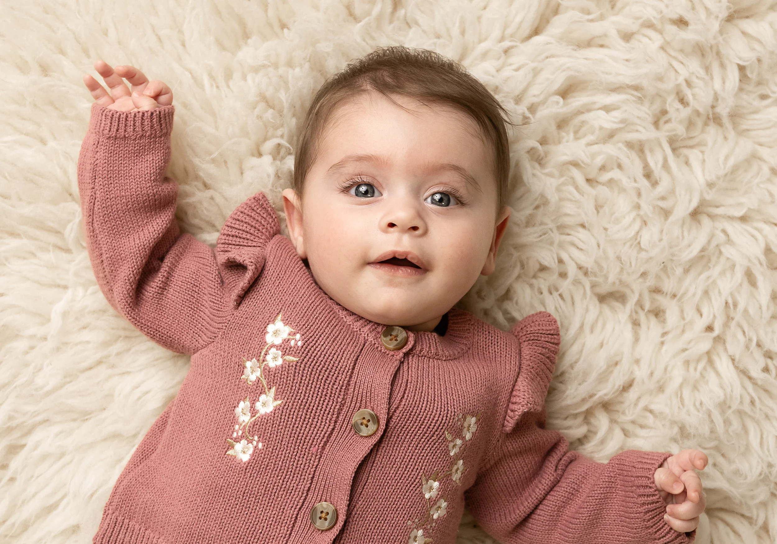 A baby girl with blue eyes and brown hair lying on a fluffy cream-colored blanket, wearing a pink knitted cardigan with floral embroidery.