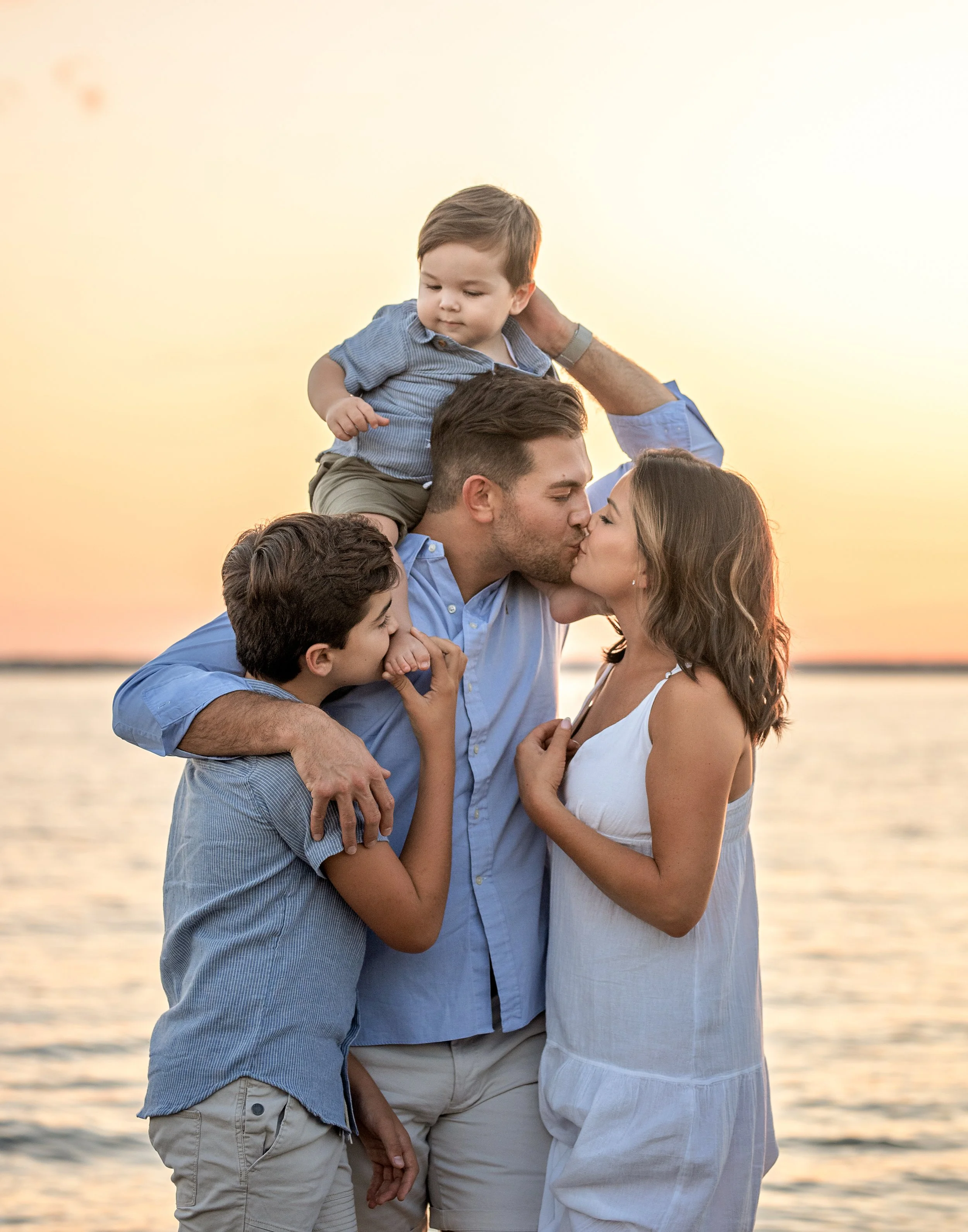 Family of four celebrating on the beach at sunset, kissing and embracing, with a young boy sitting on his father's shoulders, all dressed in light summer clothing.