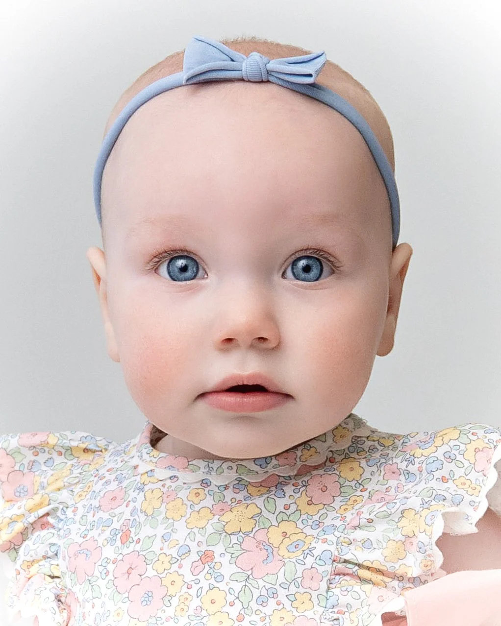 A close-up of a baby girl with blue eyes, wearing a floral dress and a light blue bow headband.