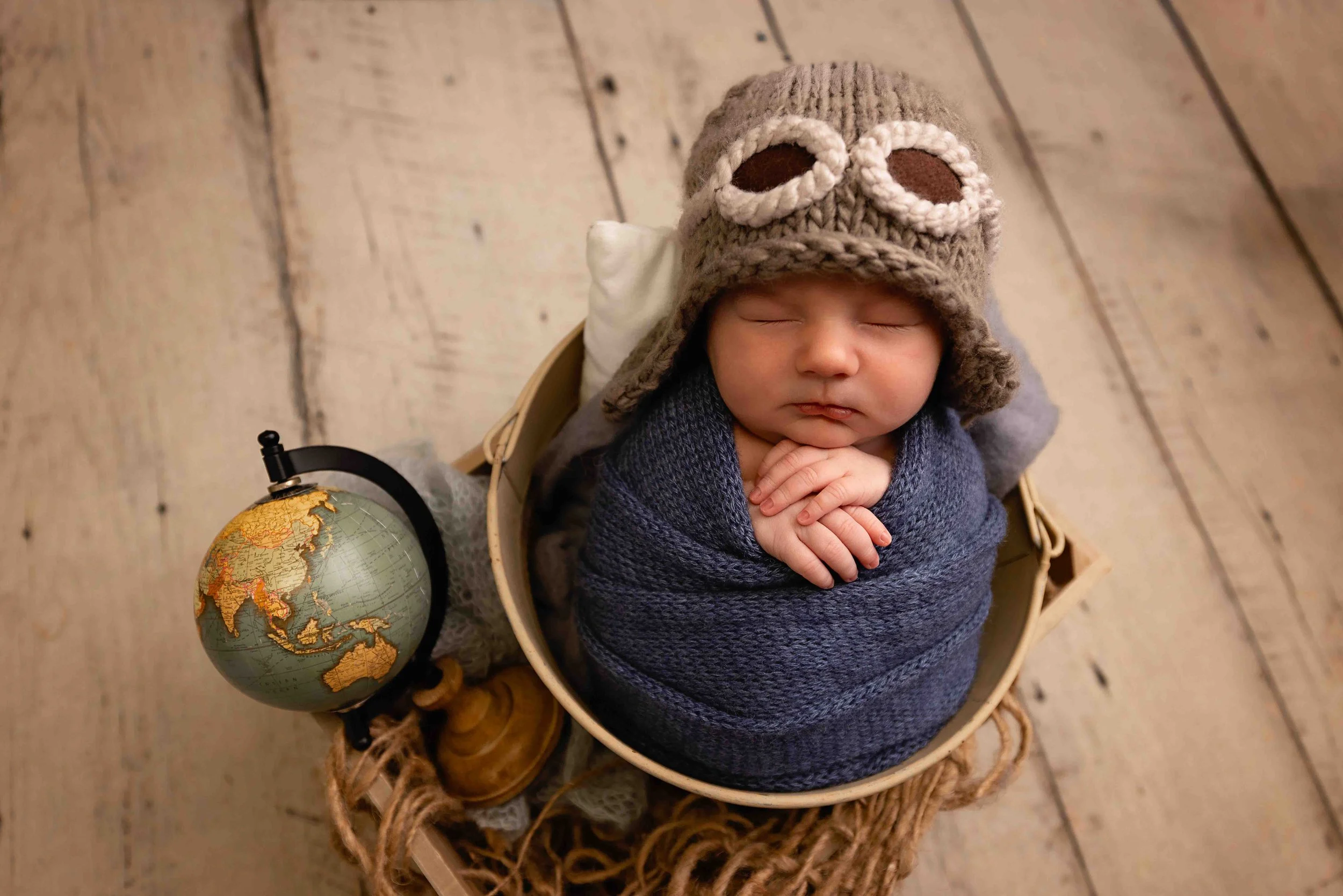 Rhode Island newborn photography swaddled baby boy sleeping in a bucket on a wood floor wearing a knit pilot hat next to a globe in studio by Anna Kay Photography
