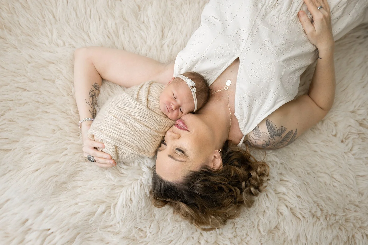 newborn baby in a cream colored swaddle laying with a mom on a textured neutral flokati rug