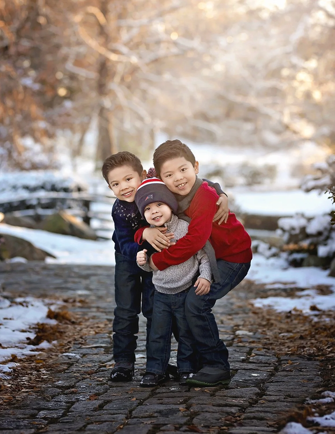 Three young boys hugging and smiling outdoors during winter, with snow on the ground and trees in the background.