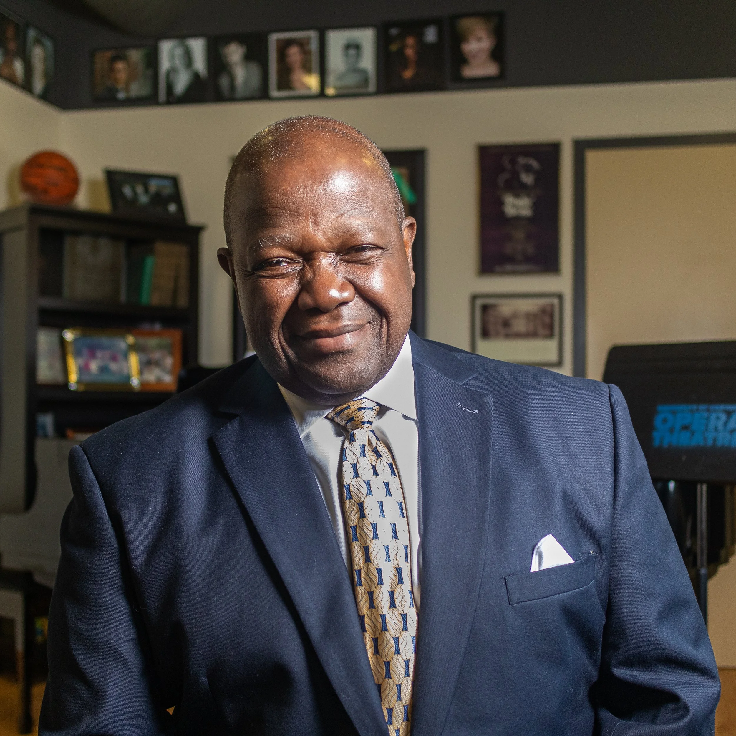 A middle-aged man in a navy suit and patterned tie smiling in an office setting with framed photos and awards on the wall behind him.