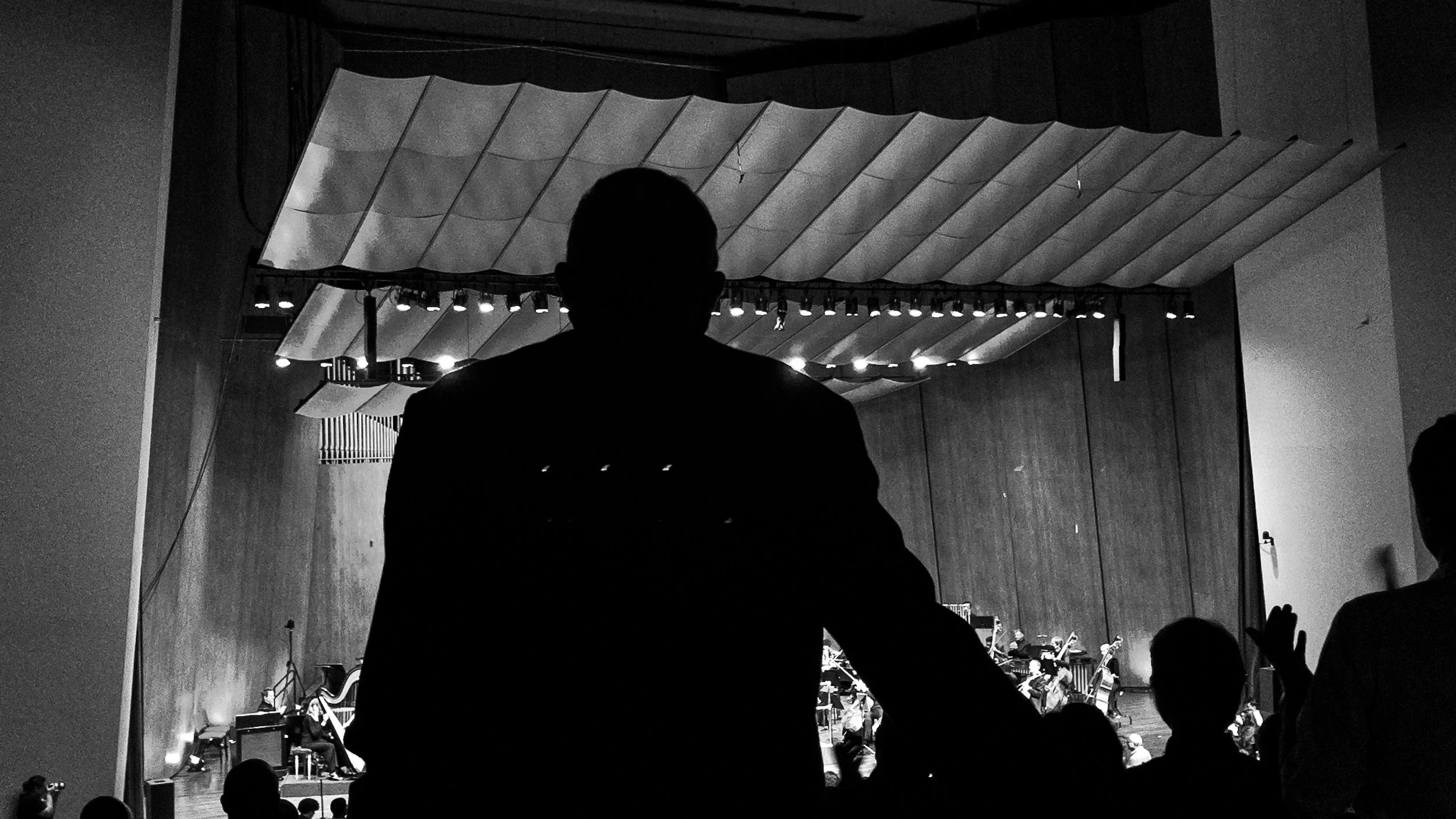 Silhouette of a person watching a performance on stage, with musicians and a conductor visible, in a concert hall with overhead acoustic panels.