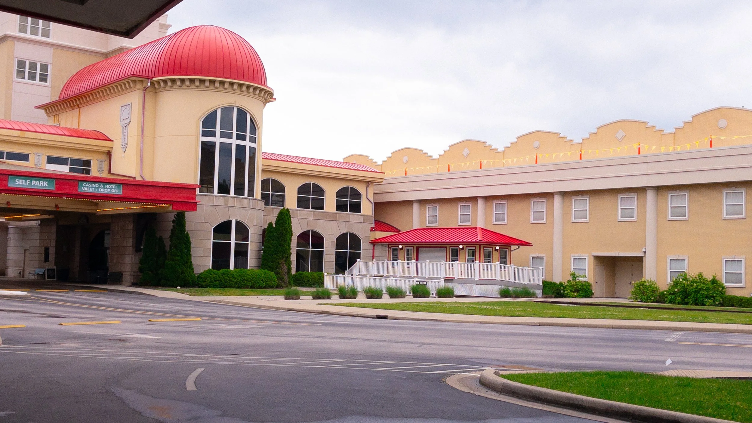 Exterior of a hotel with yellow walls and red roofs, including a circular corner tower with tall windows, a parking area, a small porch with white railing, and landscaped bushes and trees.