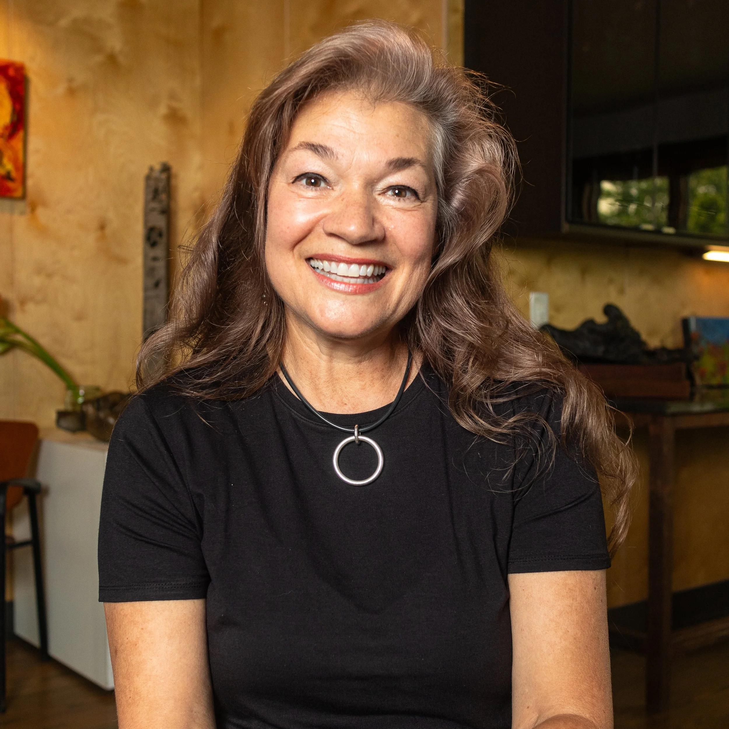 A smiling woman with long, wavy brown hair wearing a black t-shirt and a silver ring necklace, sitting in a warm, decorated indoor space.