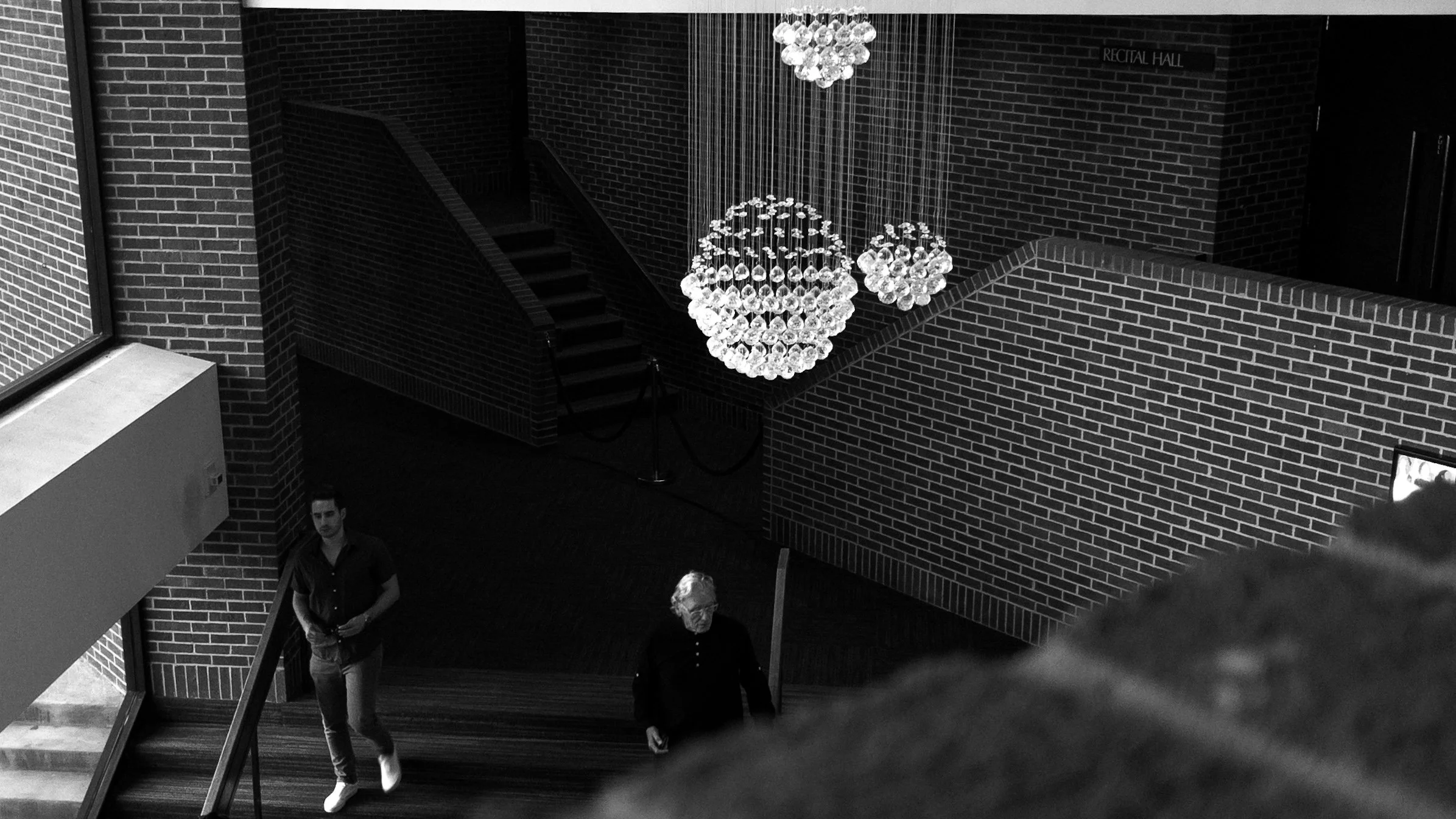 Interior of a building with brick walls, a staircase, a chandelier, and two men walking; one younger man on the left and an older man in the center.