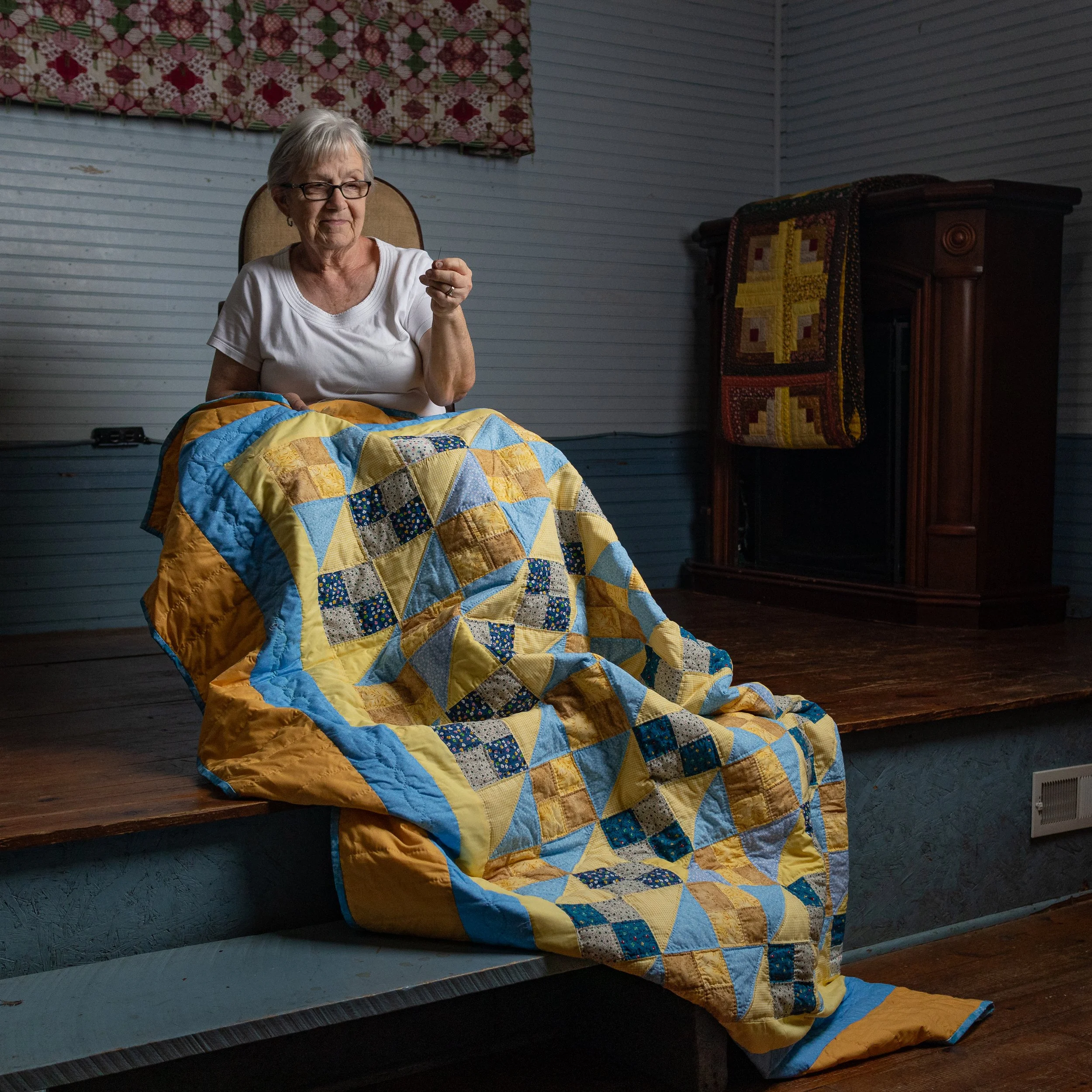 An elderly woman with short gray hair and glasses, wearing a white t-shirt, sitting on a chair with a patchwork quilt covering her lap, inside a cozy room with wood floors and vintage furniture.