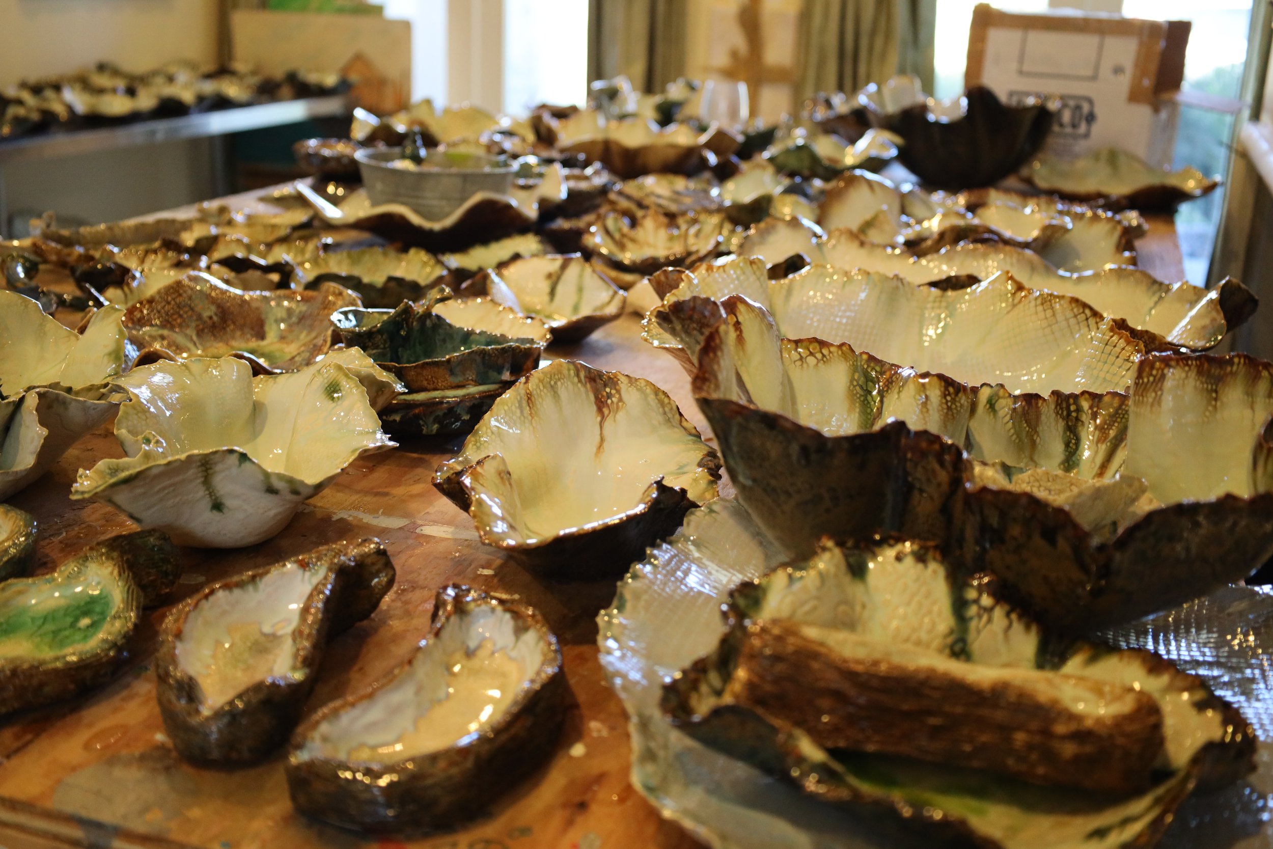 A workshop table with numerous homemade ceramic oyster shell-shaped bowls of varying sizes, glazed in earthy tones, with some stacked or scattered.