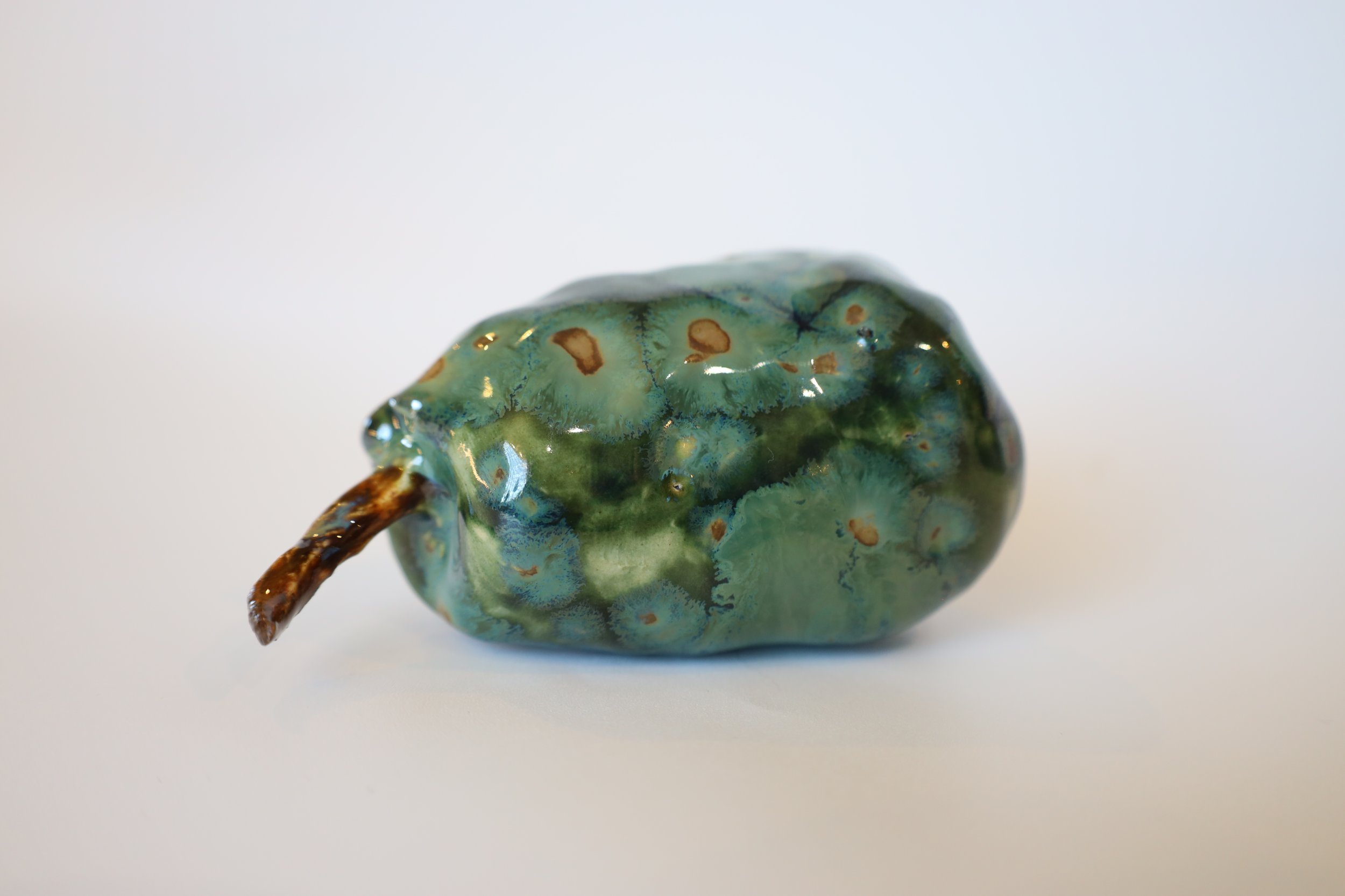 A close-up of a small, irregularly shaped glass object with a tiny brown stem, featuring a marbled green and blue pattern with brown spots, placed against a plain white background.