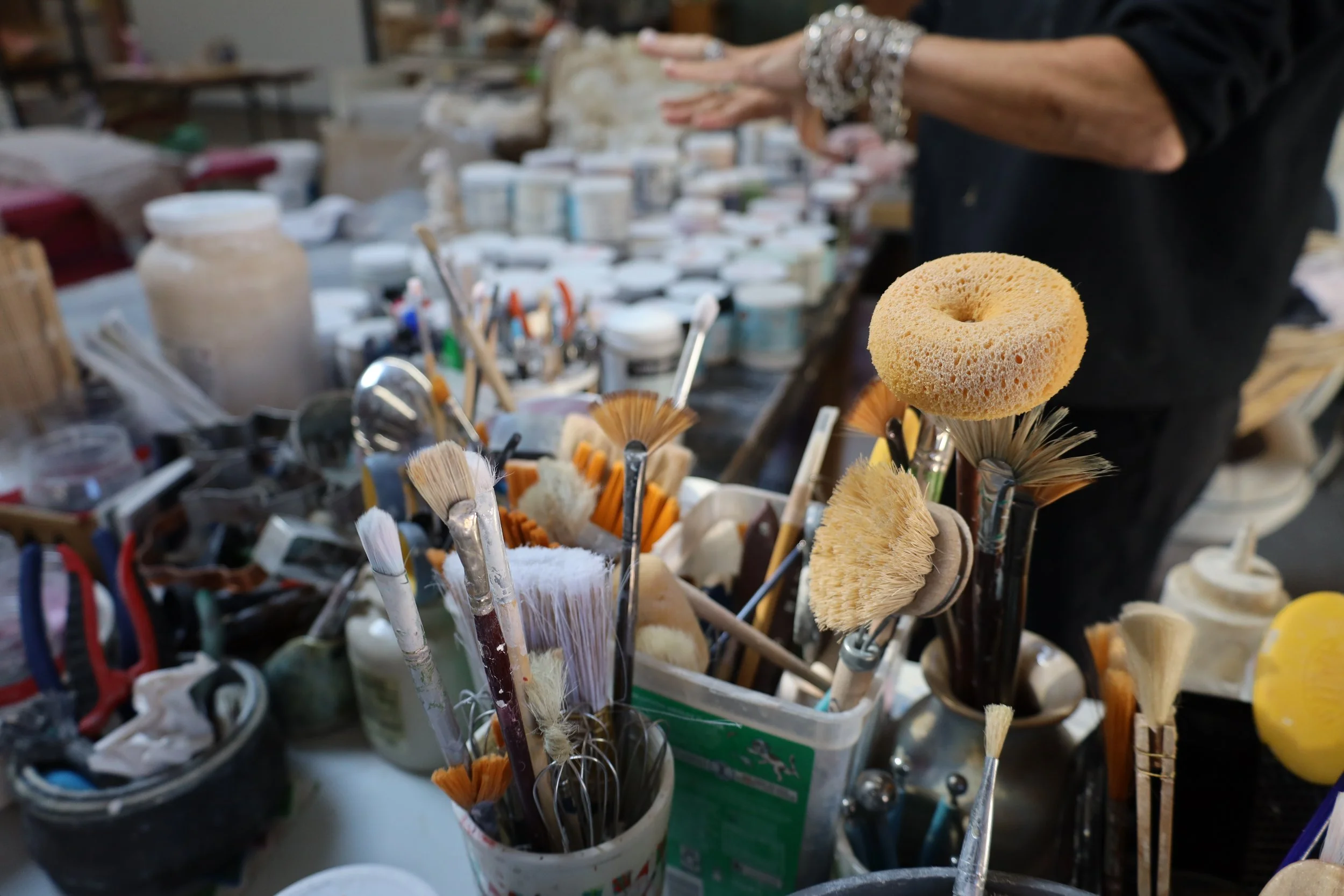 A collection of paintbrushes and a natural sea sponge in various containers on a cluttered workspace, with a person in black clothing in the background.