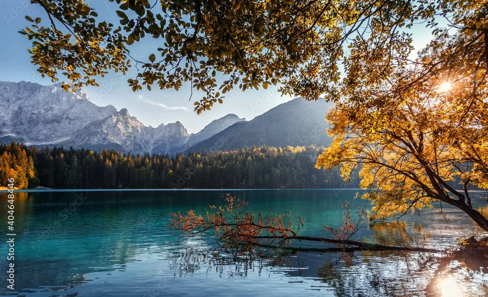 Scenic view of a lake surrounded by trees with autumn foliage, mountains in the background, and the sun shining through the branches.