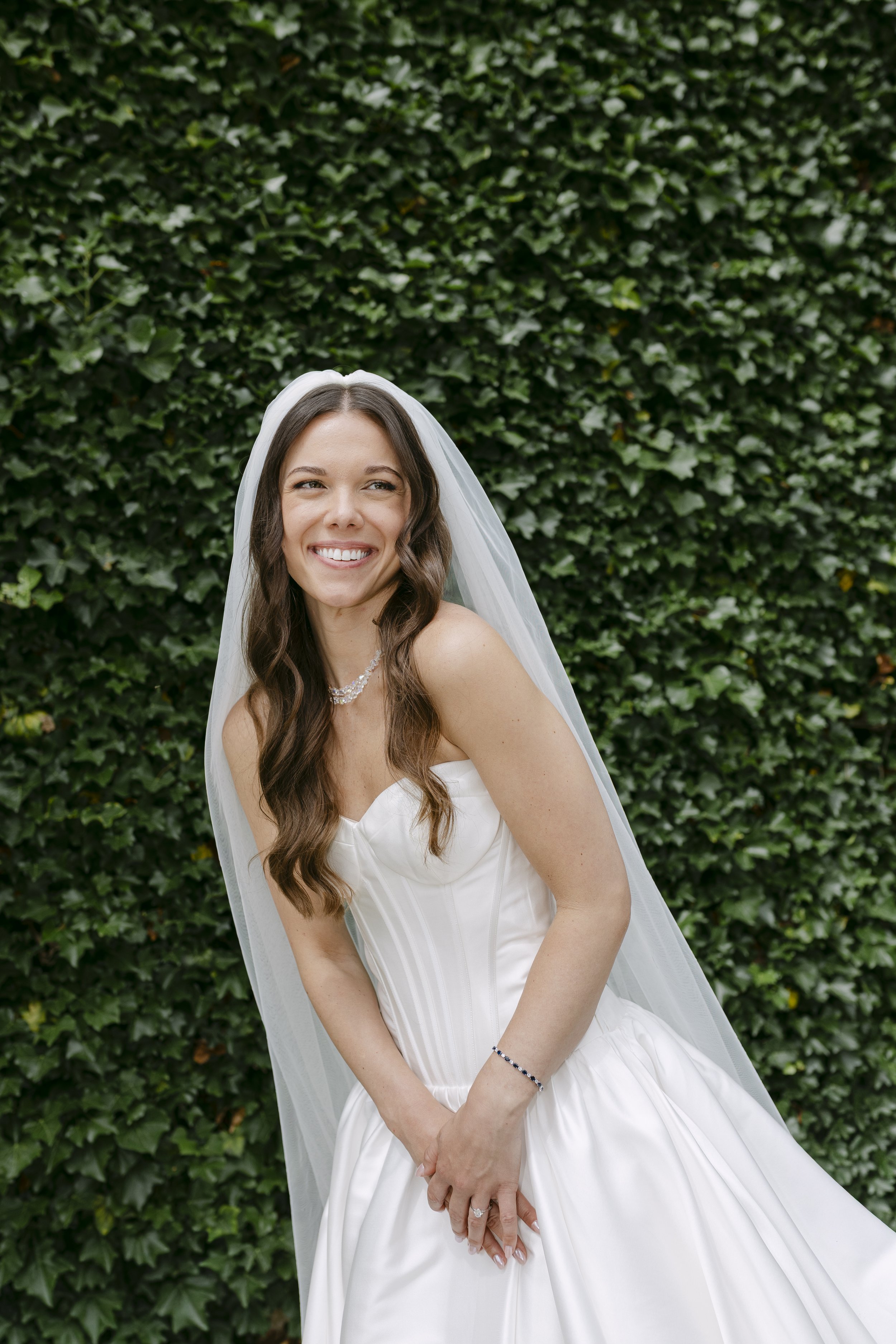 Jeune femme souriante en robe de mariée, avec voilette, devant un mur de lierre vert.