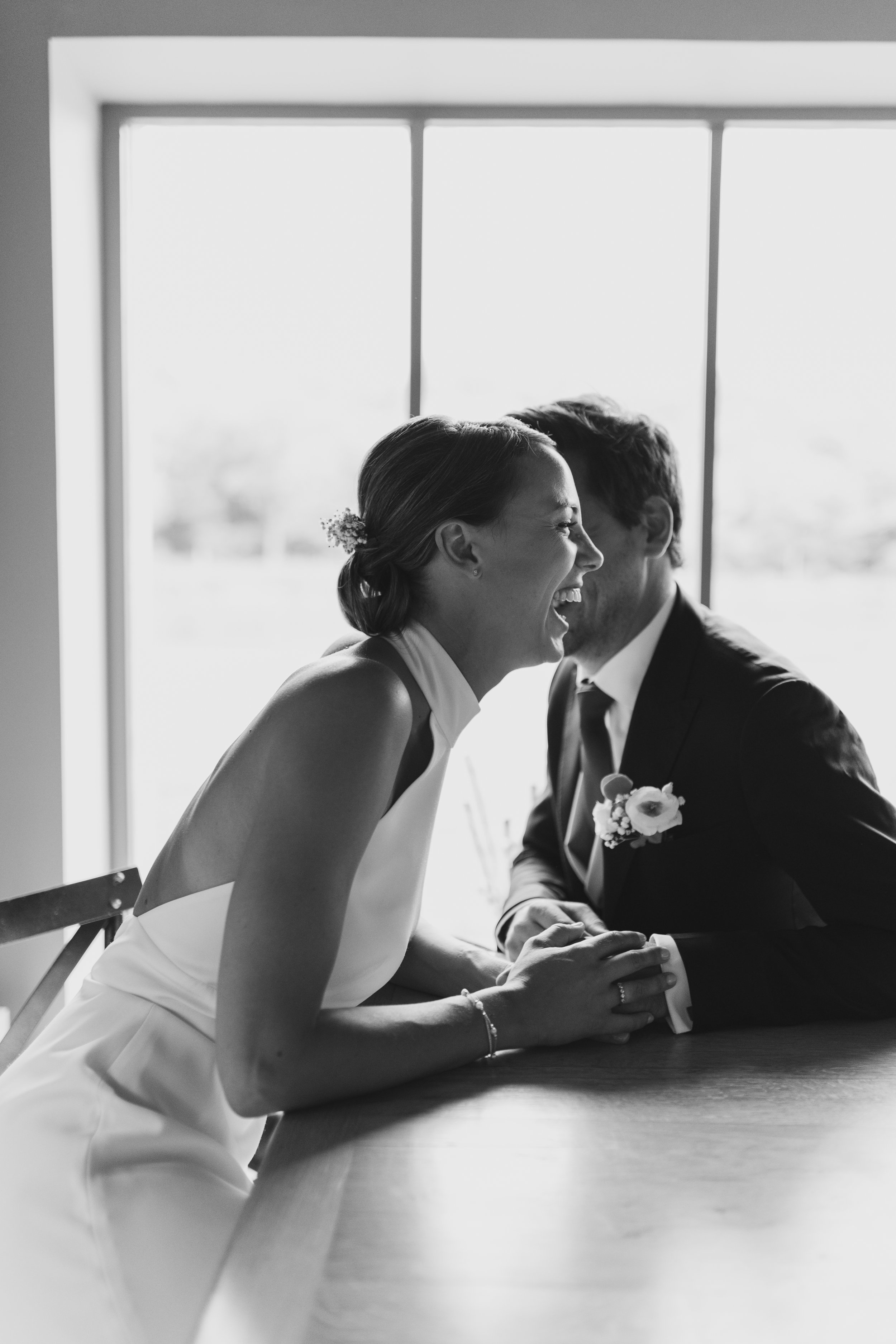 Un couple de mariés souriants, assis à une table près d'une grande fenêtre, en train de rire et de partager un moment joyeux lors de leur mariage.