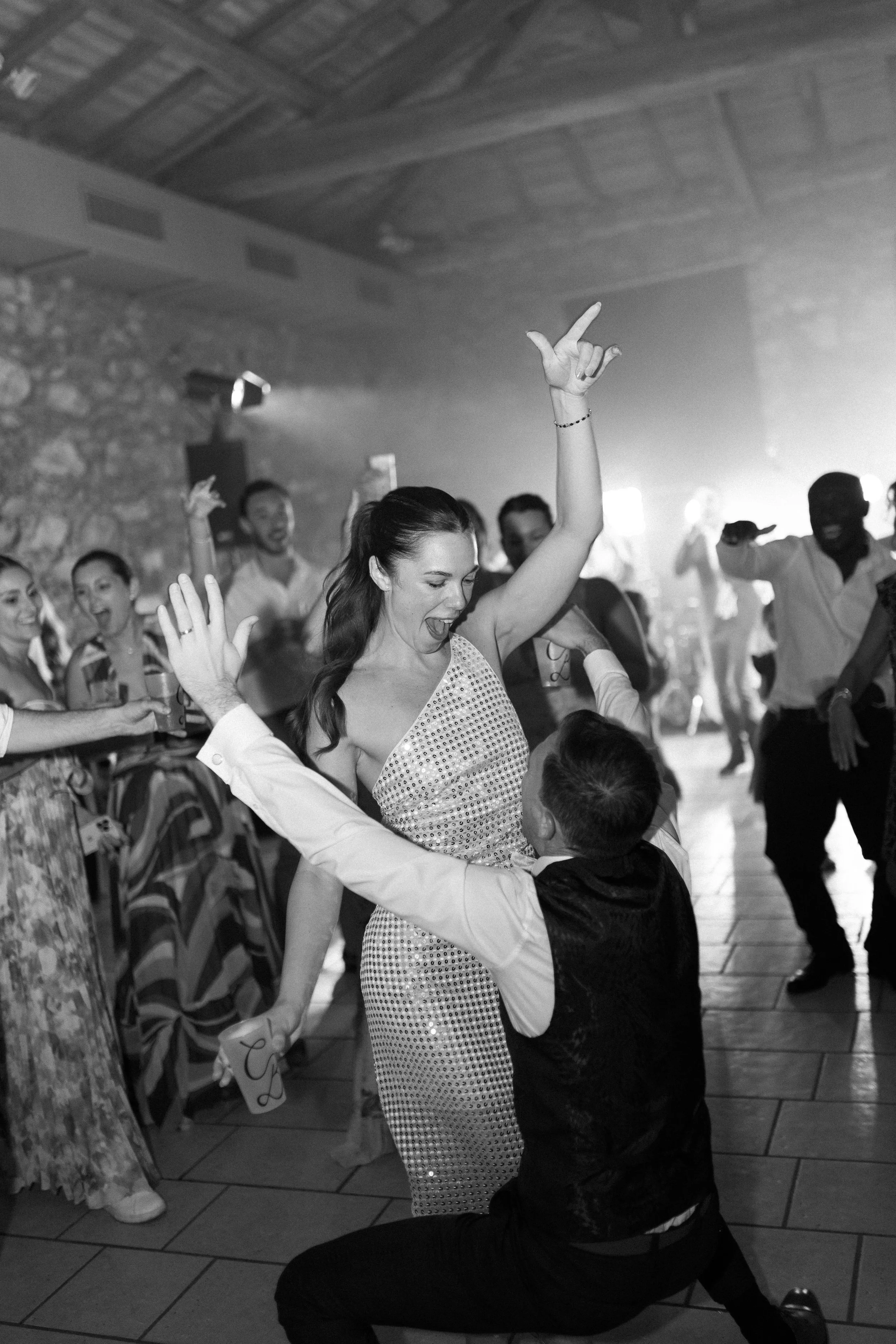 Une femme en robe brillante danse joyeusement avec un homme lors d'une fête ou d'un mariage, entourée d'autres personnes qui dansent et sourient.