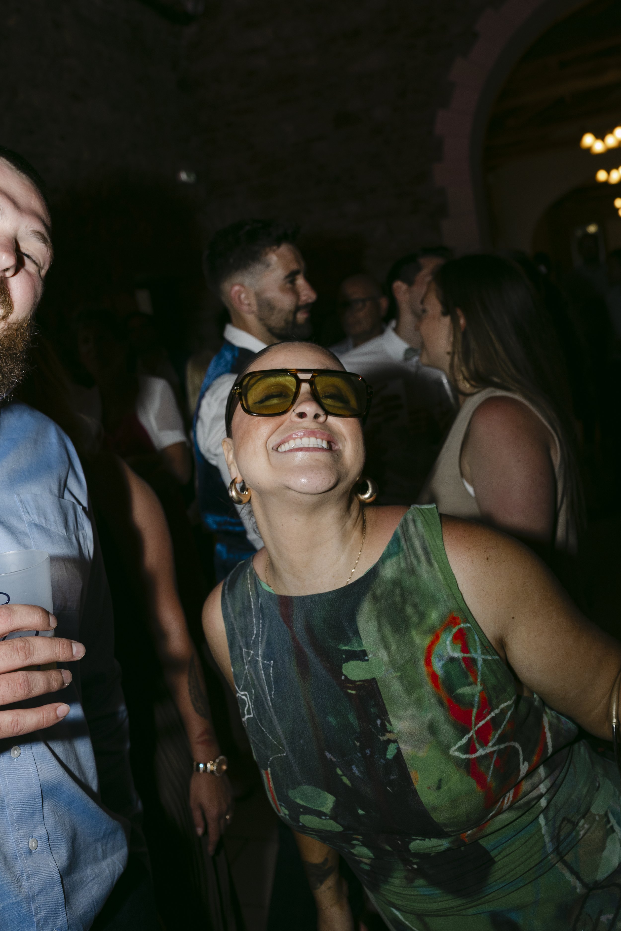 Une femme souriante portant des lunettes de soleil jaunes et une robe à motifs colorés lors d'une fête ou d'une réception, entourée d'autres personnes dans un lieu sombre.