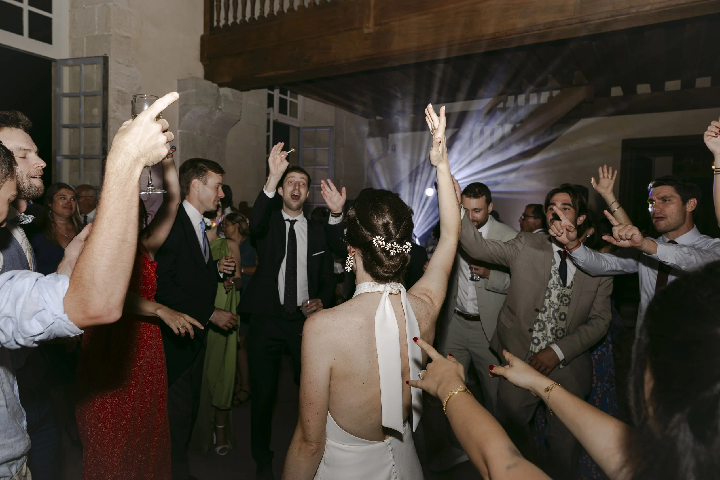 Une femme portant une robe blanche avec une décoration florale dans les cheveux danse entourée d'invités lors d'une fête, avec des personnes qui applaudissent et lèvent les mains.