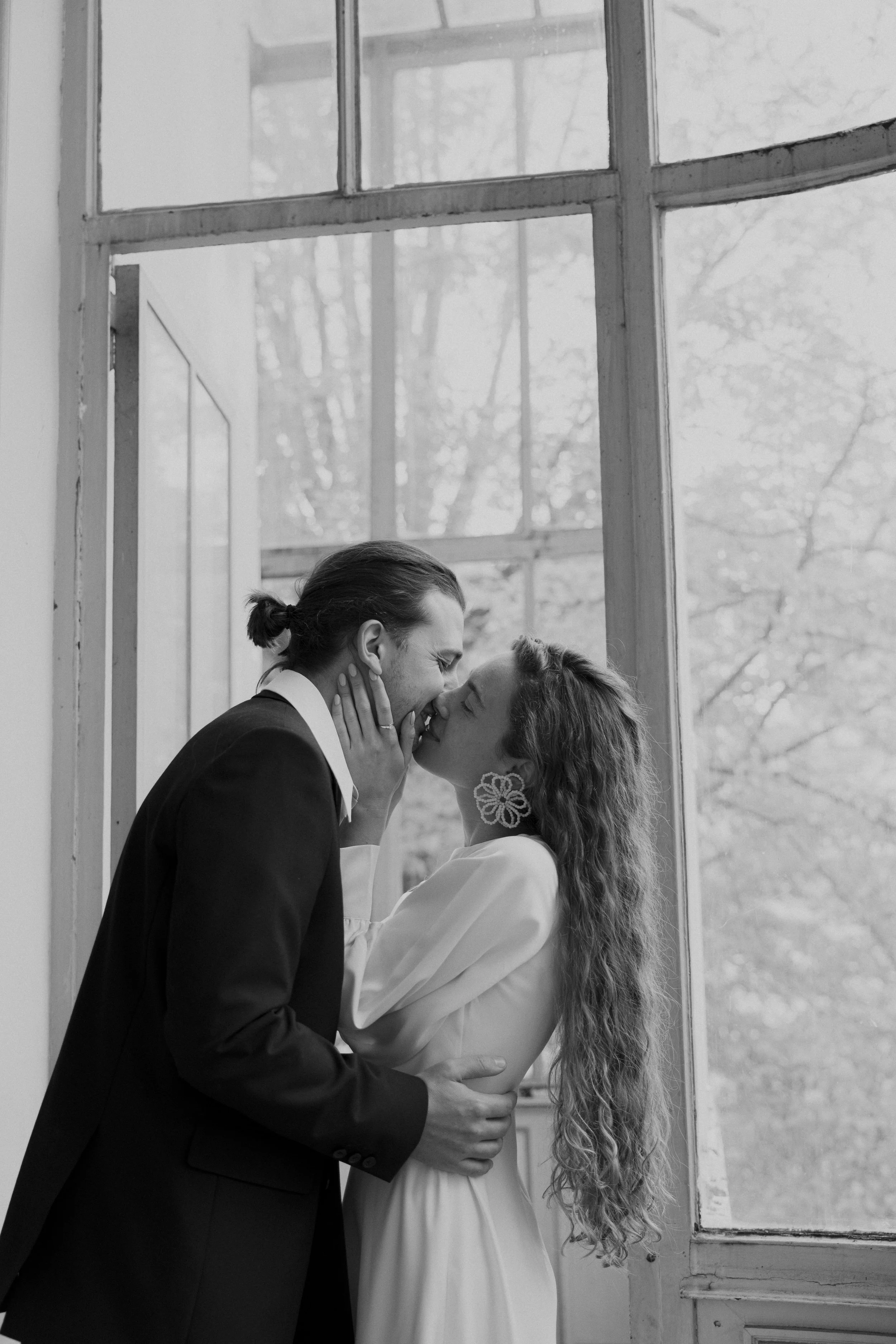 Un couple se embrasse tendrement devant une grande fenêtre, en noir et blanc.