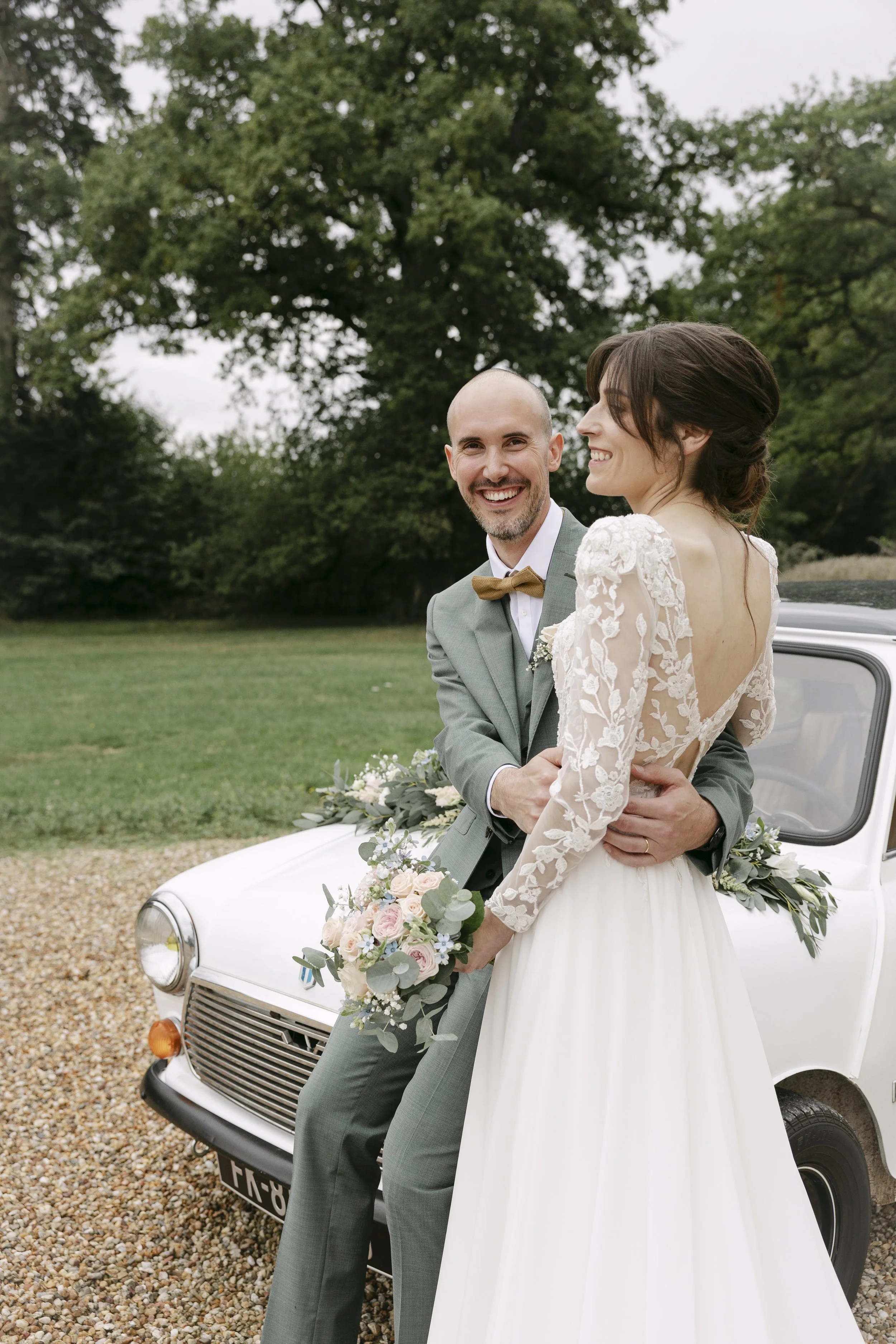 Un couple de mariés souriant en robe de mariée et costume, posant devant une petite voiture blanche, dans un cadre naturel avec des arbres.