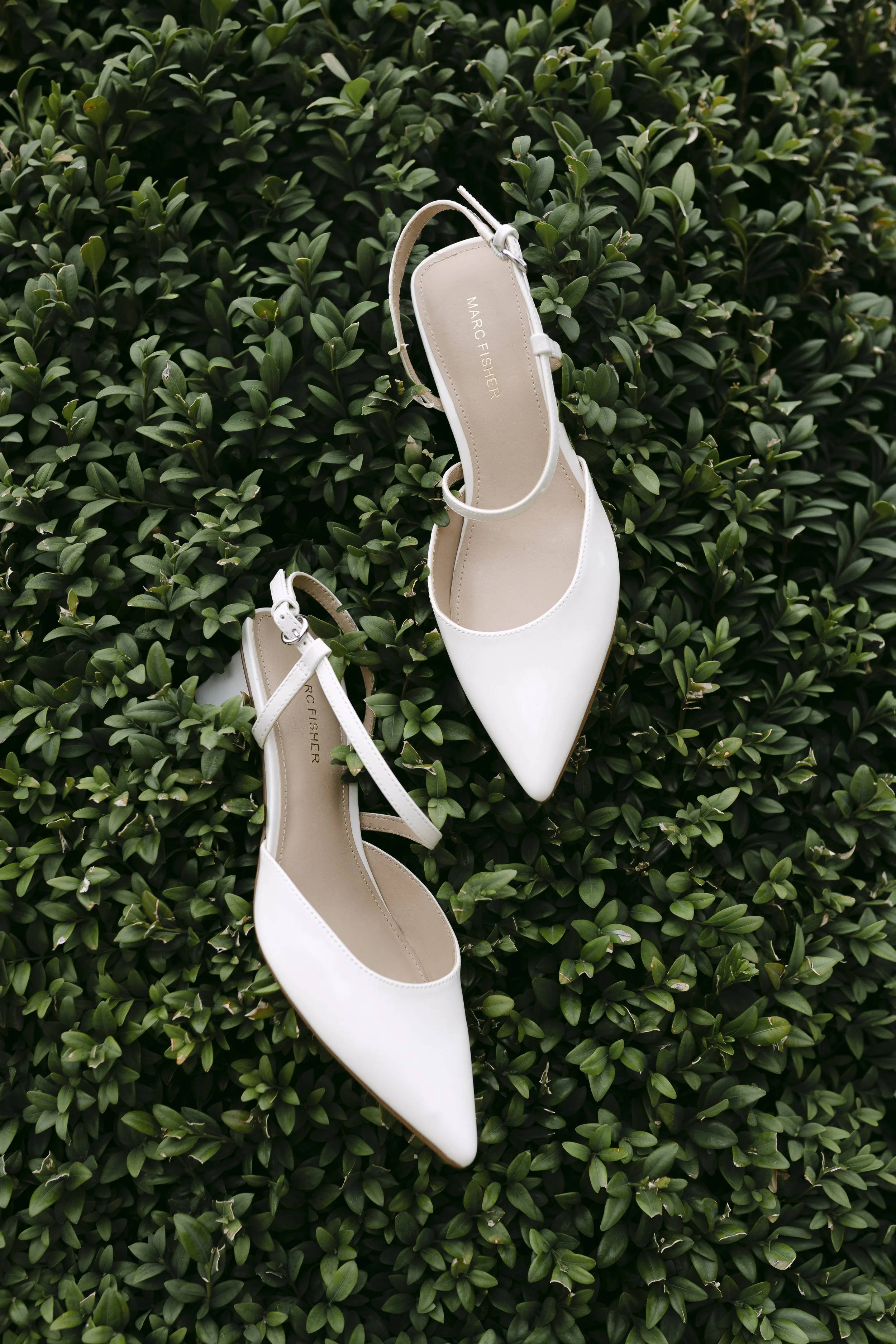 Une paire de chaussures à talons blanches en cuir brillant repose sur un tapis de feuilles vertes. La marque Marc Fisher est visible à l'intérieur des chaussures.