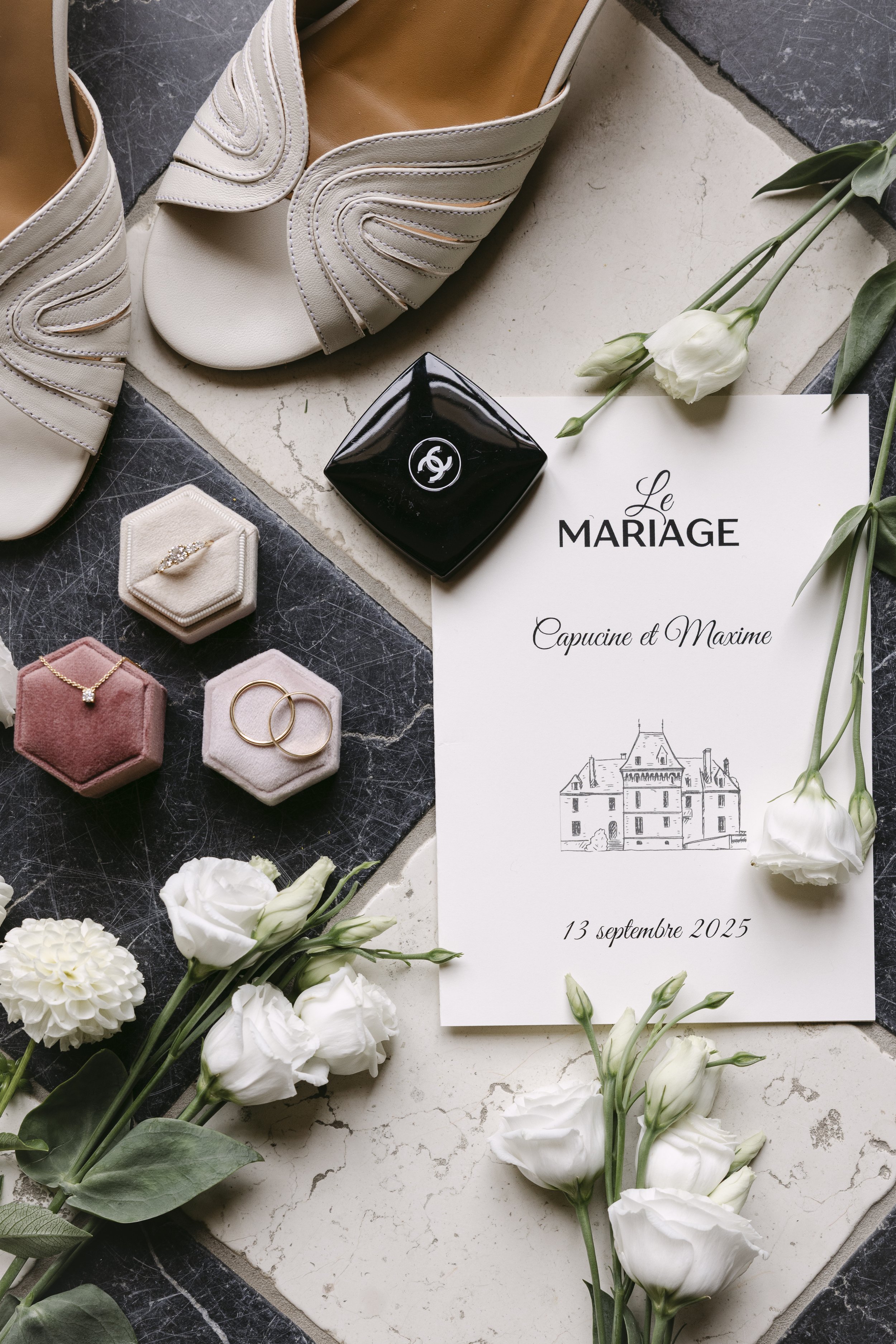 Invitation de mariage avec des fleurs blanches, chaussures beiges, bijoux en or et en diamant, un ruban noir avec un logo, et un marque-page.