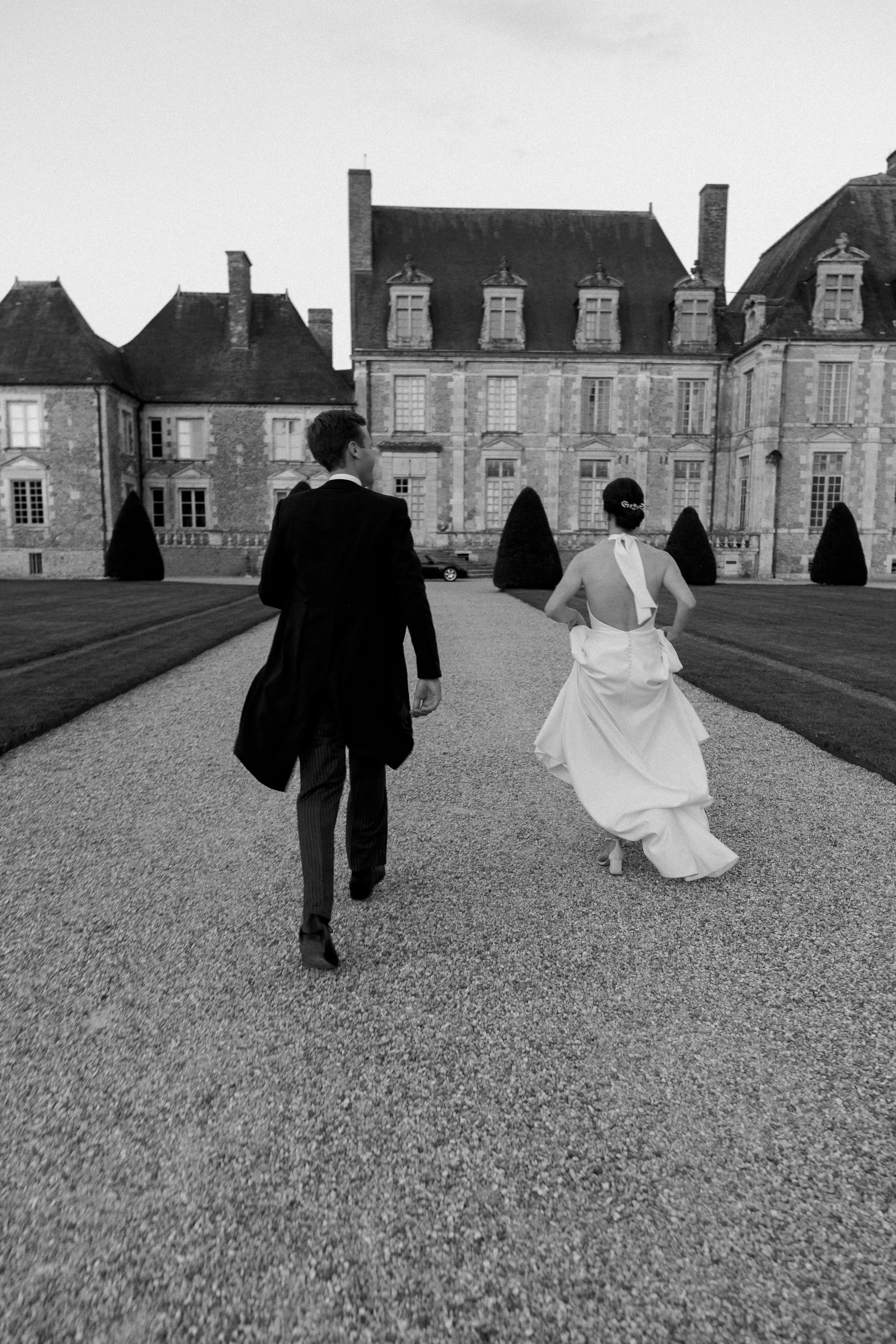 Un couple en robe de mariage et costume marche devant un grand château en pierre, en noir et blanc.